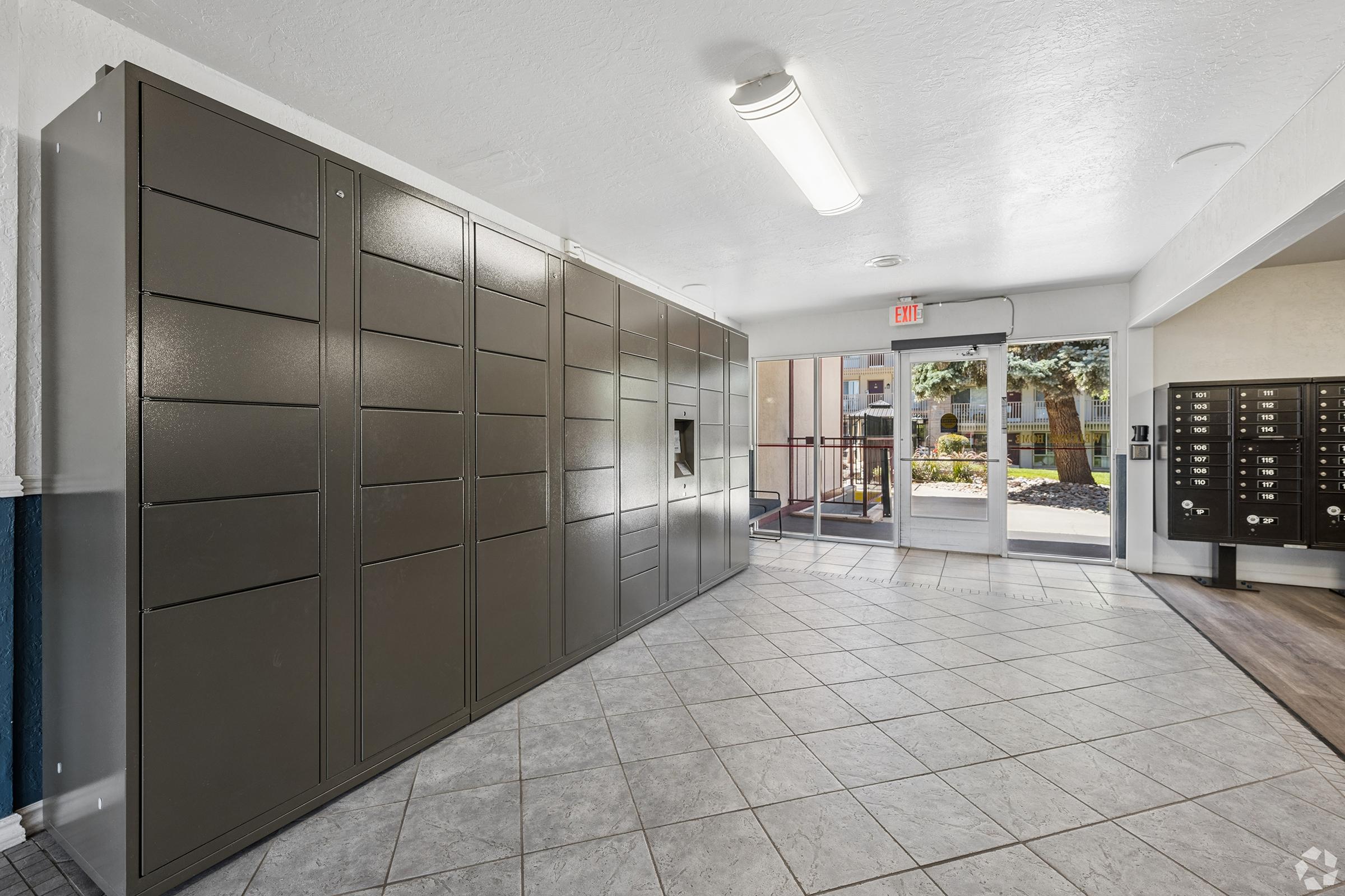 A spacious indoor area featuring multiple gray mail lockers on the left side and a glass door leading outside. Large windows allow natural light to fill the space, which has tiled flooring and a clean, modern design, suitable for receiving packages or mail.