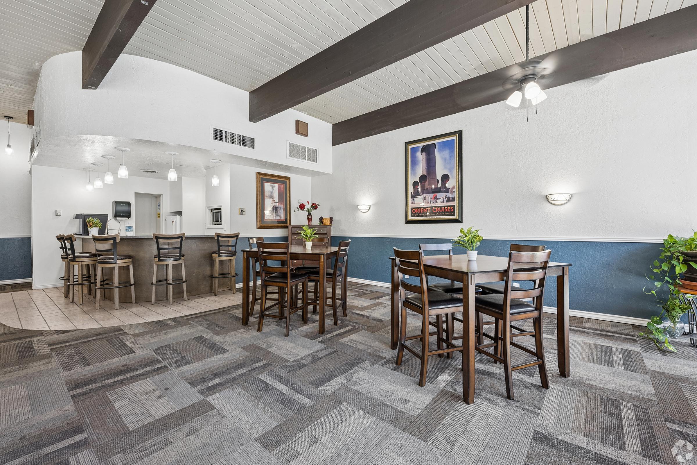 Dining area featuring several wooden tables and chairs, with a light-colored ceiling and dark beams. The walls are painted in contrasting colors, and there are decorative elements like plants and artwork. Soft lighting fixtures hang from the ceiling, enhancing the inviting atmosphere.