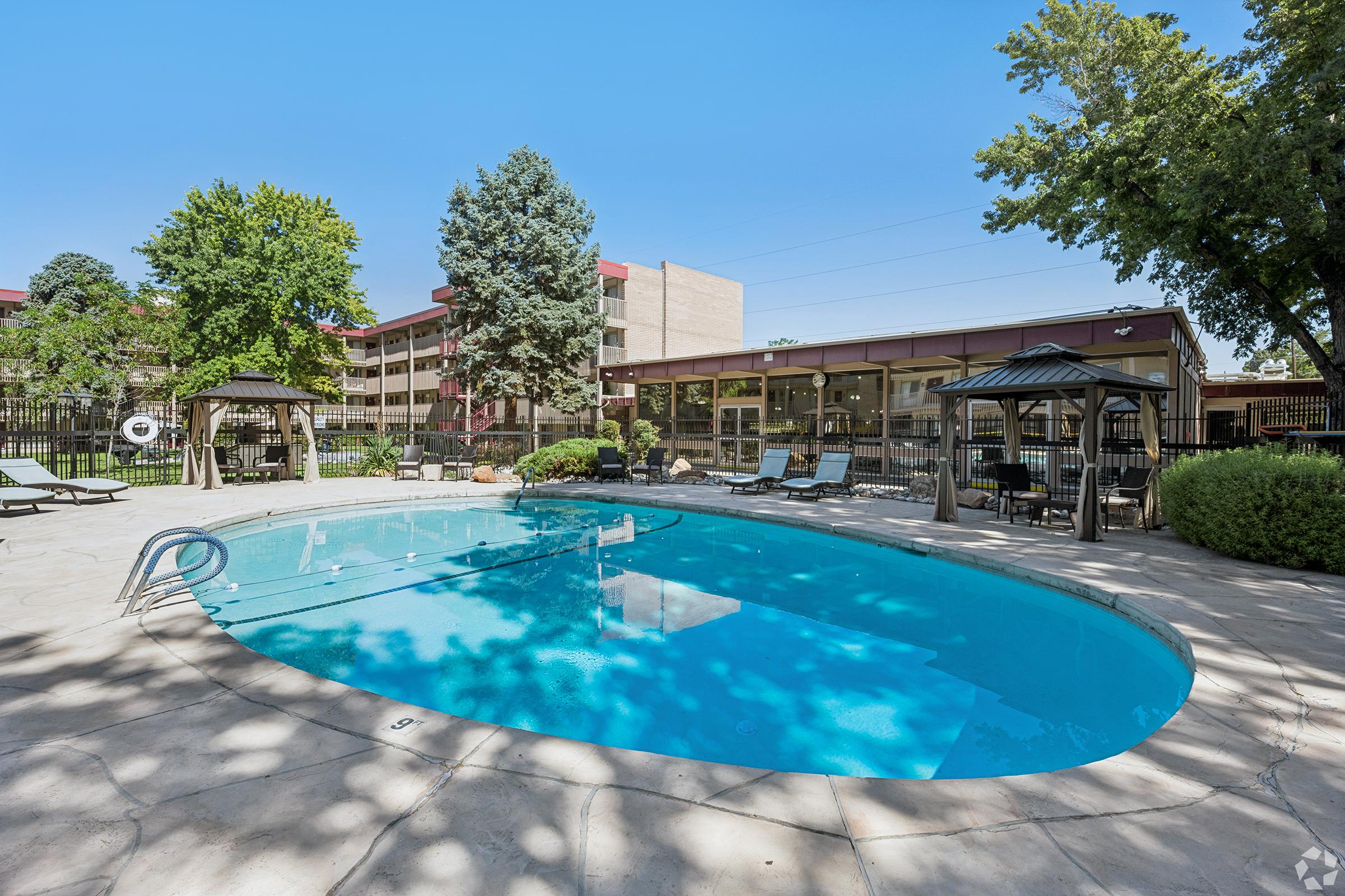A clear blue swimming pool surrounded by lounge chairs and shaded by trees. In the background, there is a building with large windows, along with a gazebo area for relaxation. The scene is bright and sunny, creating an inviting atmosphere for leisure and relaxation.