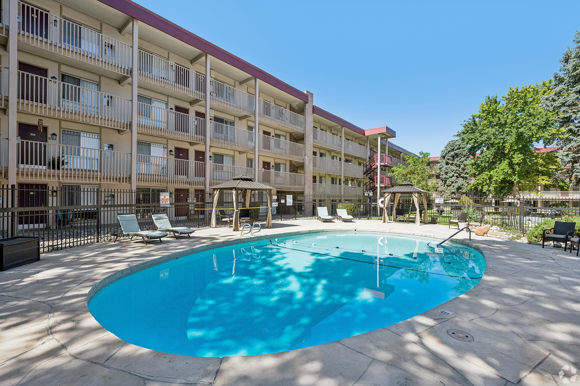A tranquil swimming pool surrounded by lounge chairs and shaded seating areas in a landscaped courtyard of a multi-story building. The clear blue sky offers a bright atmosphere, while trees provide natural shade around the pool area.