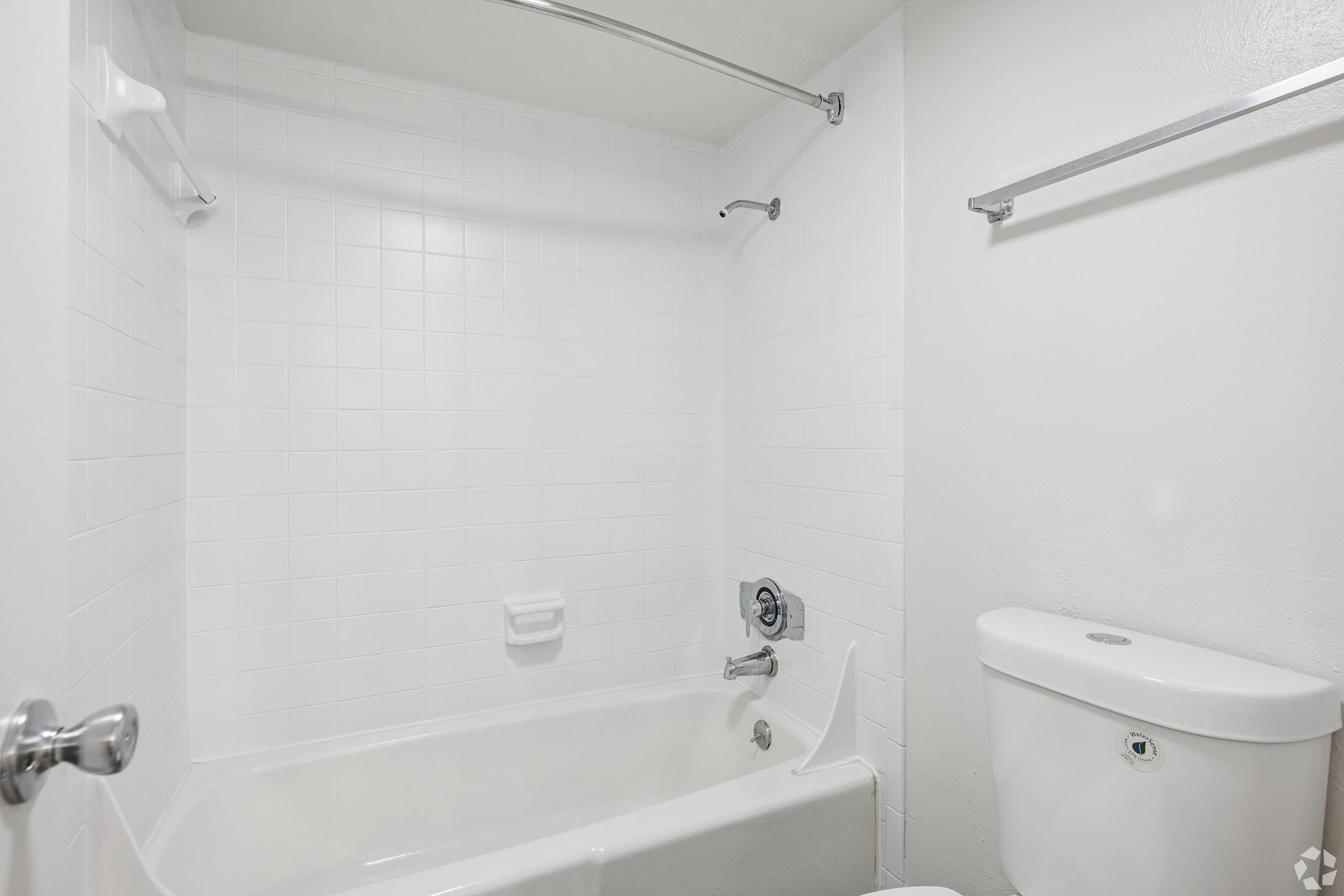 A clean, modern bathroom featuring a white tiled shower area with a bathtub, a chrome faucet, and a beige wall-mounted soap dish. The space includes a white toilet on the right side, with a simple silver towel rail above the bathtub and a bright, minimalistic design.