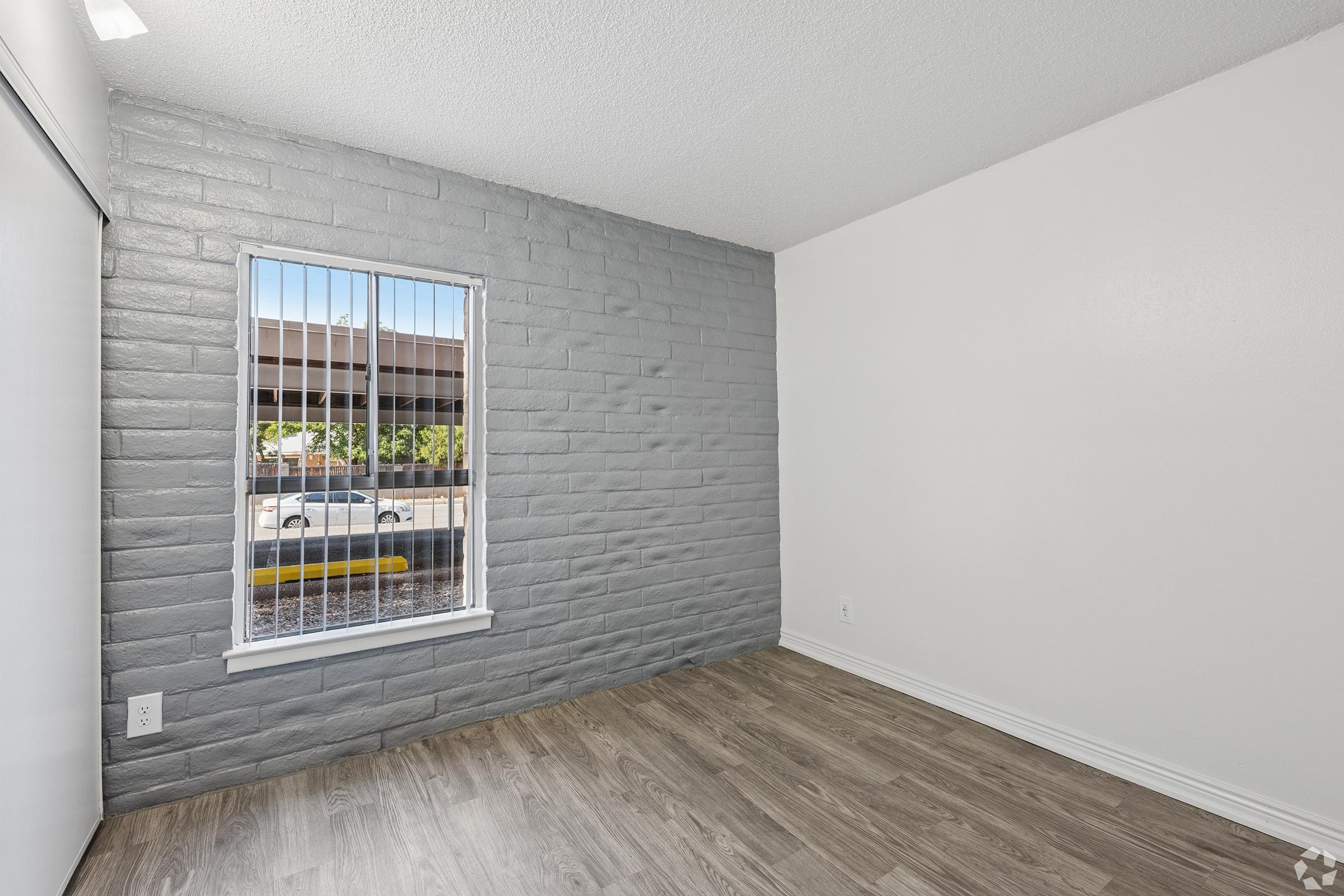 A small room featuring a gray accent wall, a window with vertical blinds allowing natural light, and light wood flooring. The wall opposite the window is painted white, creating a bright and airy atmosphere. The room is empty, showcasing its spaciousness and potential for furnishing.