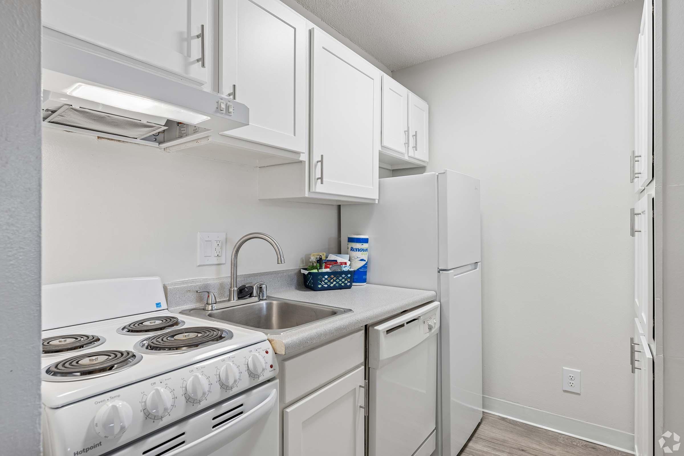 A small kitchen featuring white cabinetry, a stainless steel sink, a refrigerator, and a stove with an oven. The countertop is light gray, and there are various kitchen items, including cleaning supplies, on display. The walls are painted a neutral color, creating a bright and tidy atmosphere.