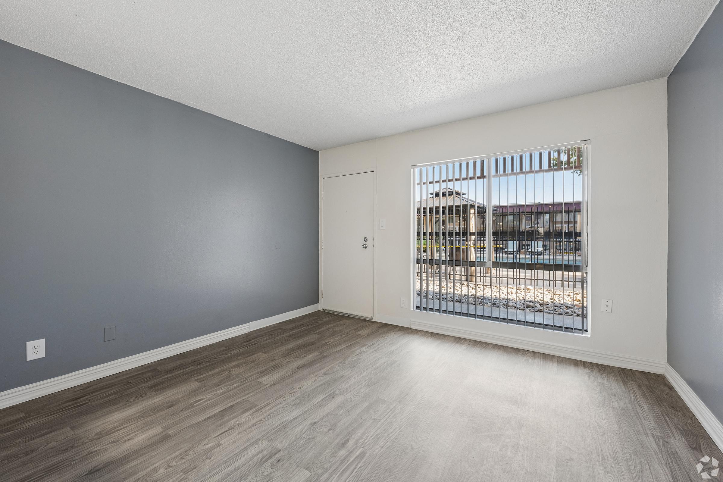 A vacant room featuring light gray walls and laminate flooring. A large window with vertical blinds lets in natural light and offers a view of an outdoor area. The entrance door is visible on the left side of the image.
