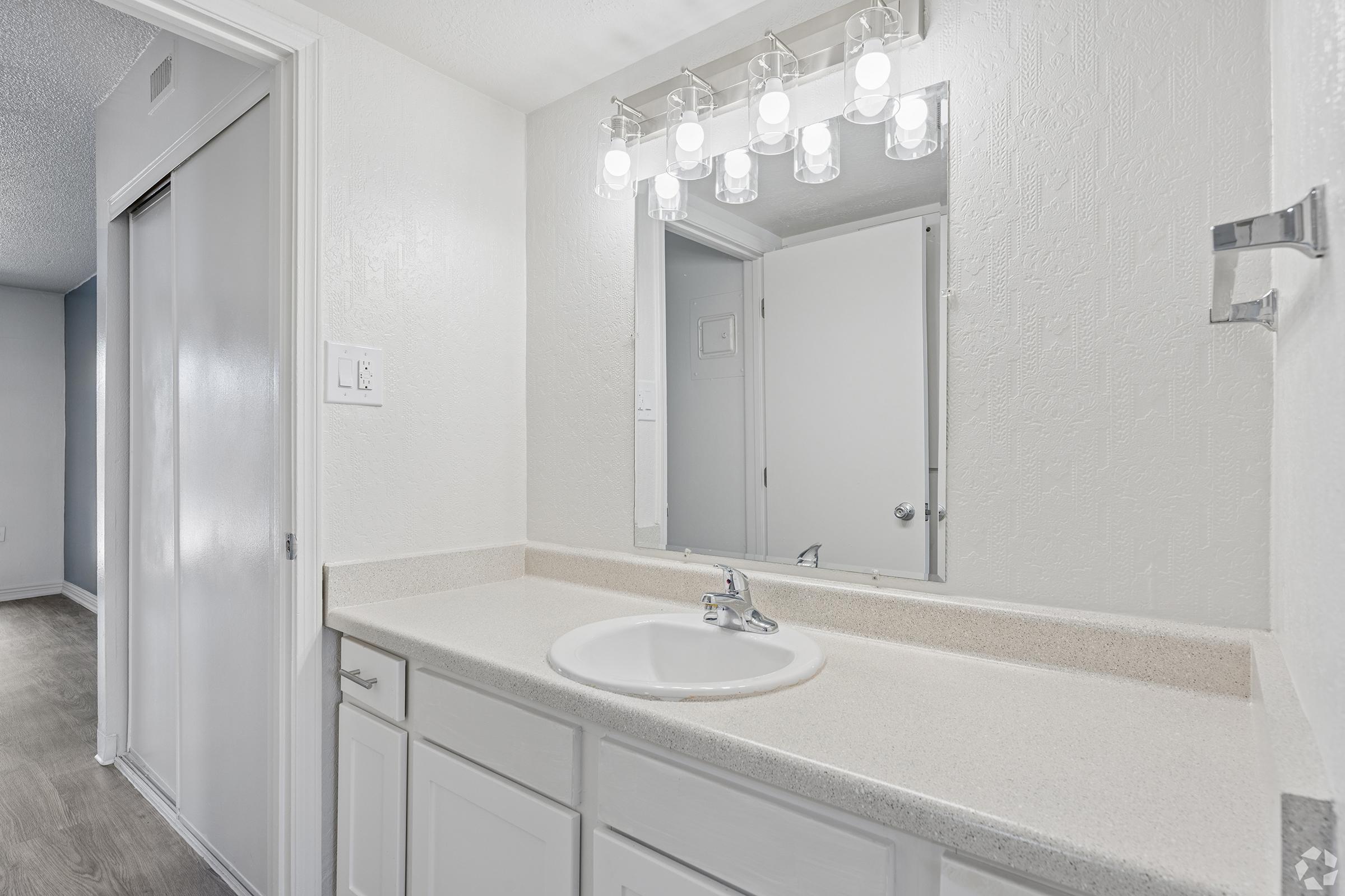 A well-lit bathroom with a clean, white vanity and a round sink. Above the sink, there are five light fixtures providing ample illumination. A large mirror is mounted on the wall, reflecting the bright space. Light-colored walls and cabinetry create a fresh and modern atmosphere. A glimpse of a hallway is visible in the background.