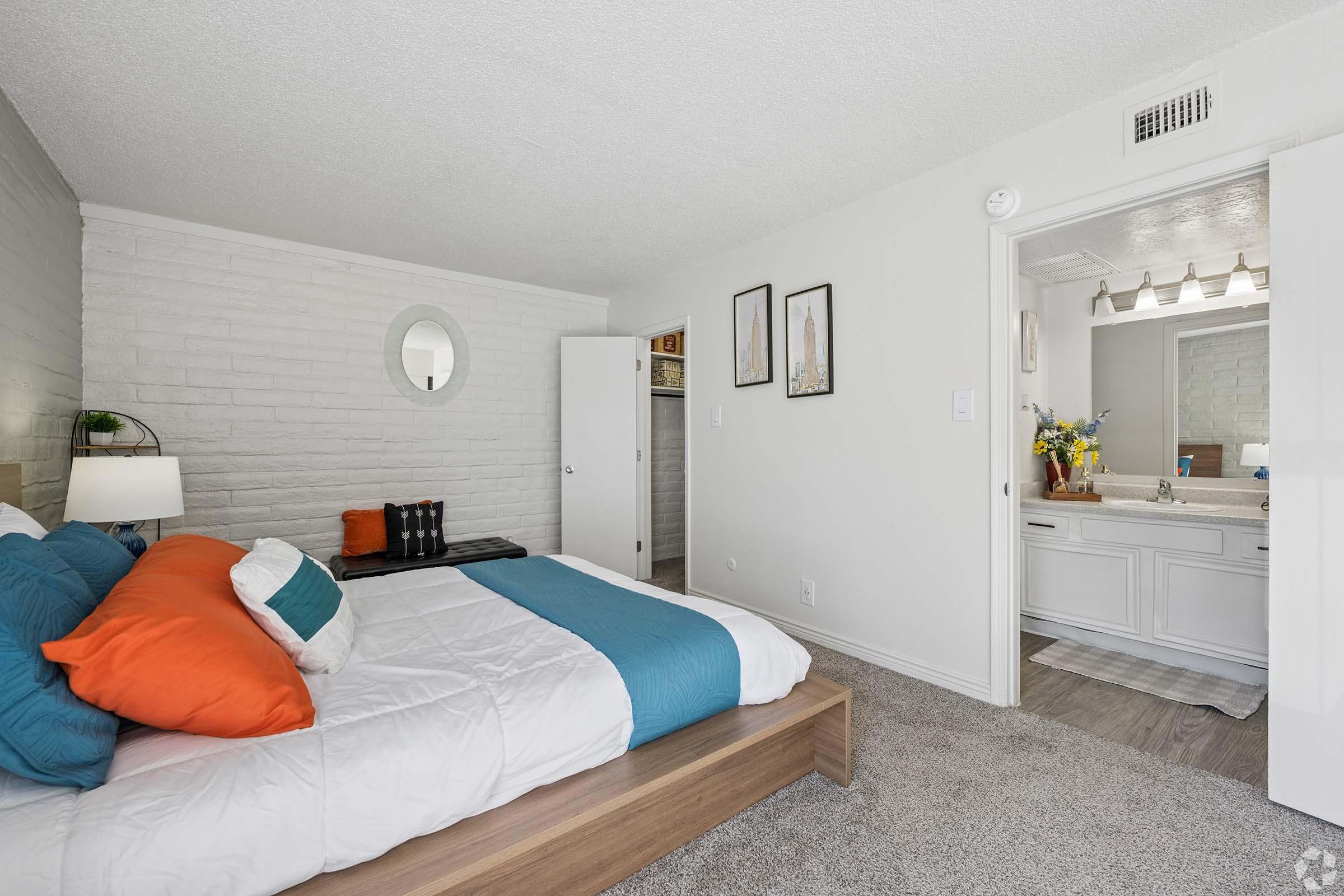 A cozy bedroom featuring a bed with blue and orange pillows, a neutral-colored comforter, and a round mirror above a nightstand. A closet is visible in the background, and a doorway leads to a bathroom adorned with vanity and floral arrangements. The walls have a light texture, and the flooring is carpeted.