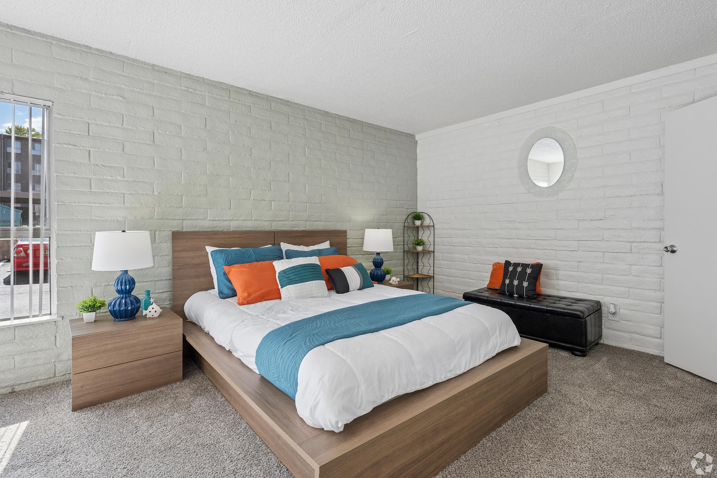 A cozy bedroom featuring a queen-sized bed with white and turquoise bedding. There are decorative pillows in orange and blue. A bedside table with blue lamps on either side, a round wall mirror, and a small black bench add charm. Light-colored brick walls and carpet enhance the inviting atmosphere.