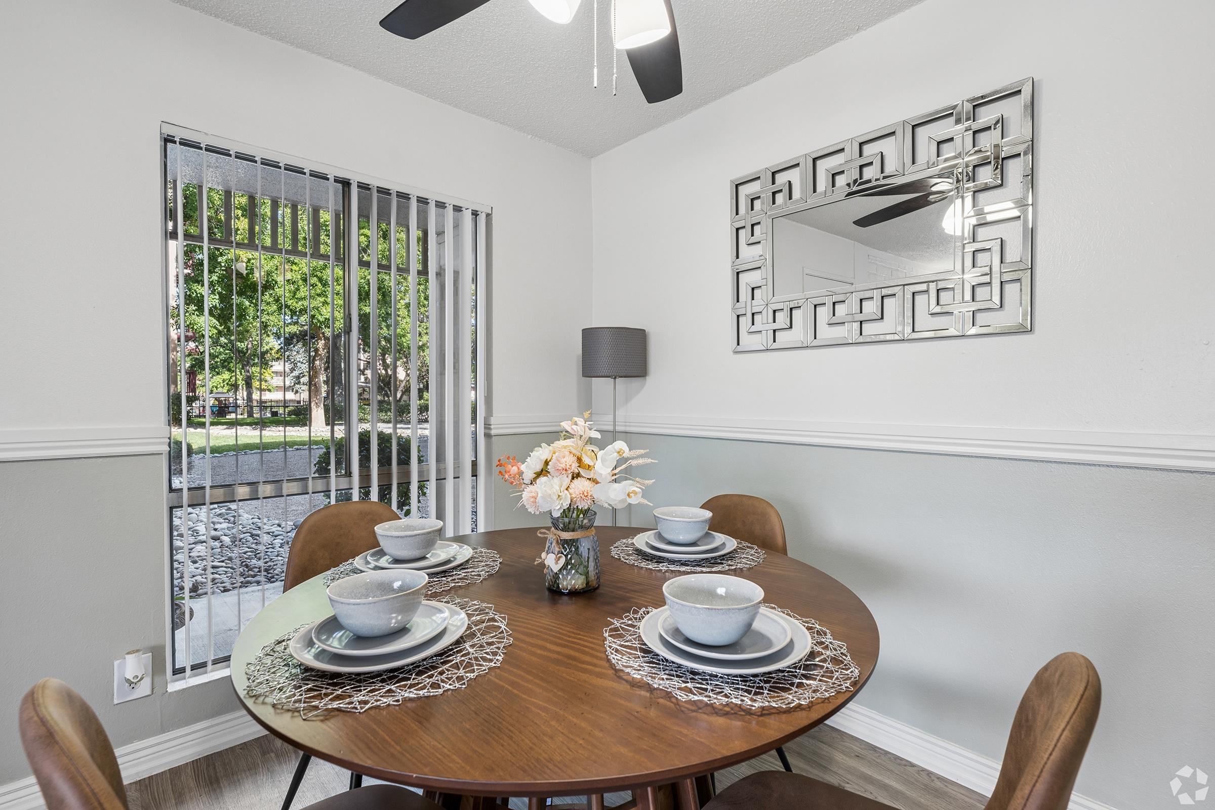 A cozy dining area featuring a round wooden table set for four with plates and decorative placemats. A vase of flowers adds color to the table. Behind, there are windows with vertical blinds, and a decorative mirror on the wall reflects natural light, enhancing the inviting ambiance.