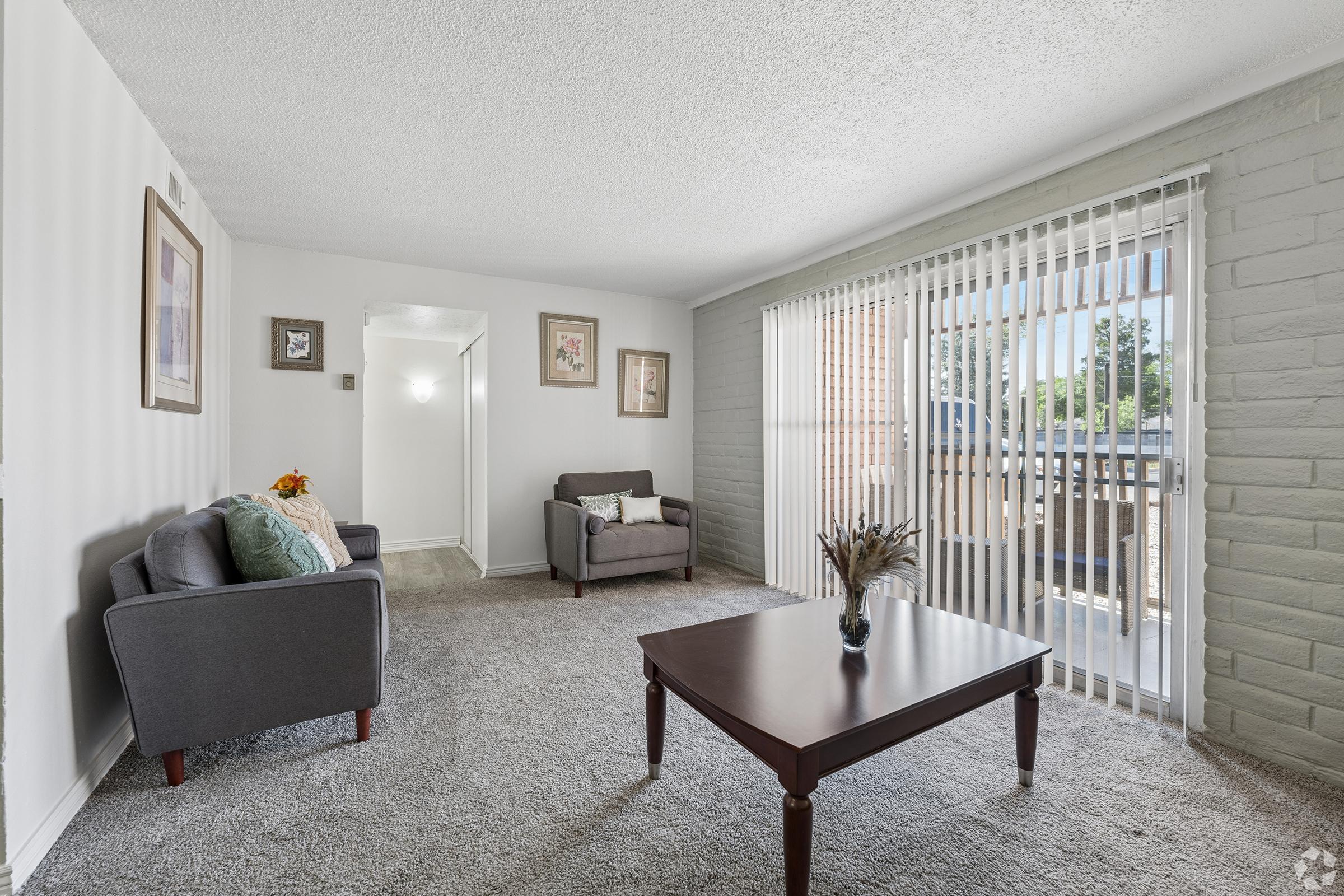 A cozy living room featuring two gray armchairs, a wooden coffee table with a vase of dried flowers, and a light-colored carpet. Large sliding glass doors allow ample natural light and provide a view of an outdoor space. The walls are painted white, with framed art pieces hanging on them.