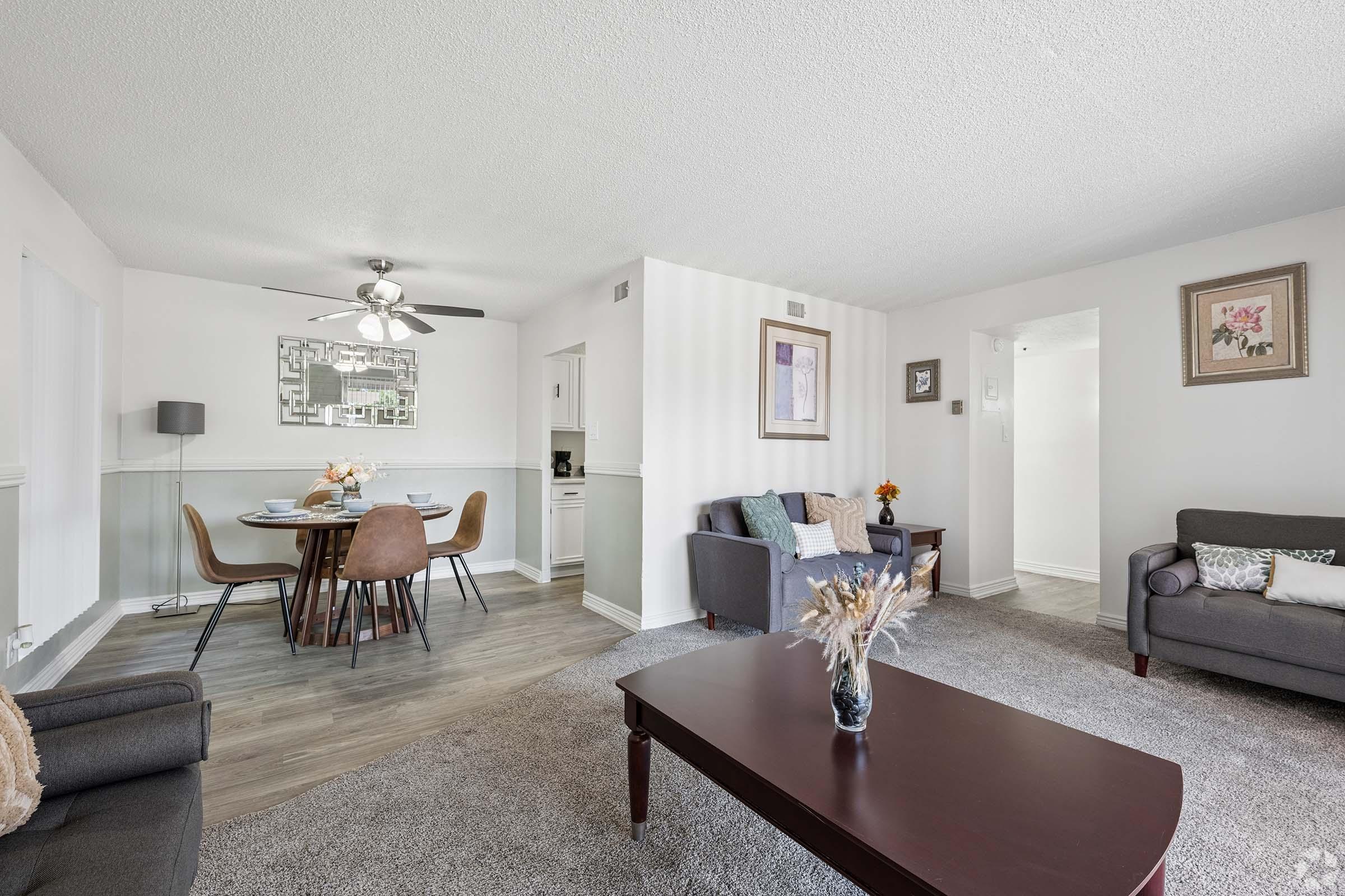 A cozy living room with two gray sofas and a coffee table on a plush carpet. A dining area with a round table and chairs is visible in the background, next to a modern kitchen. The walls are painted light, and artwork adds a touch of decor, creating a warm, inviting atmosphere.