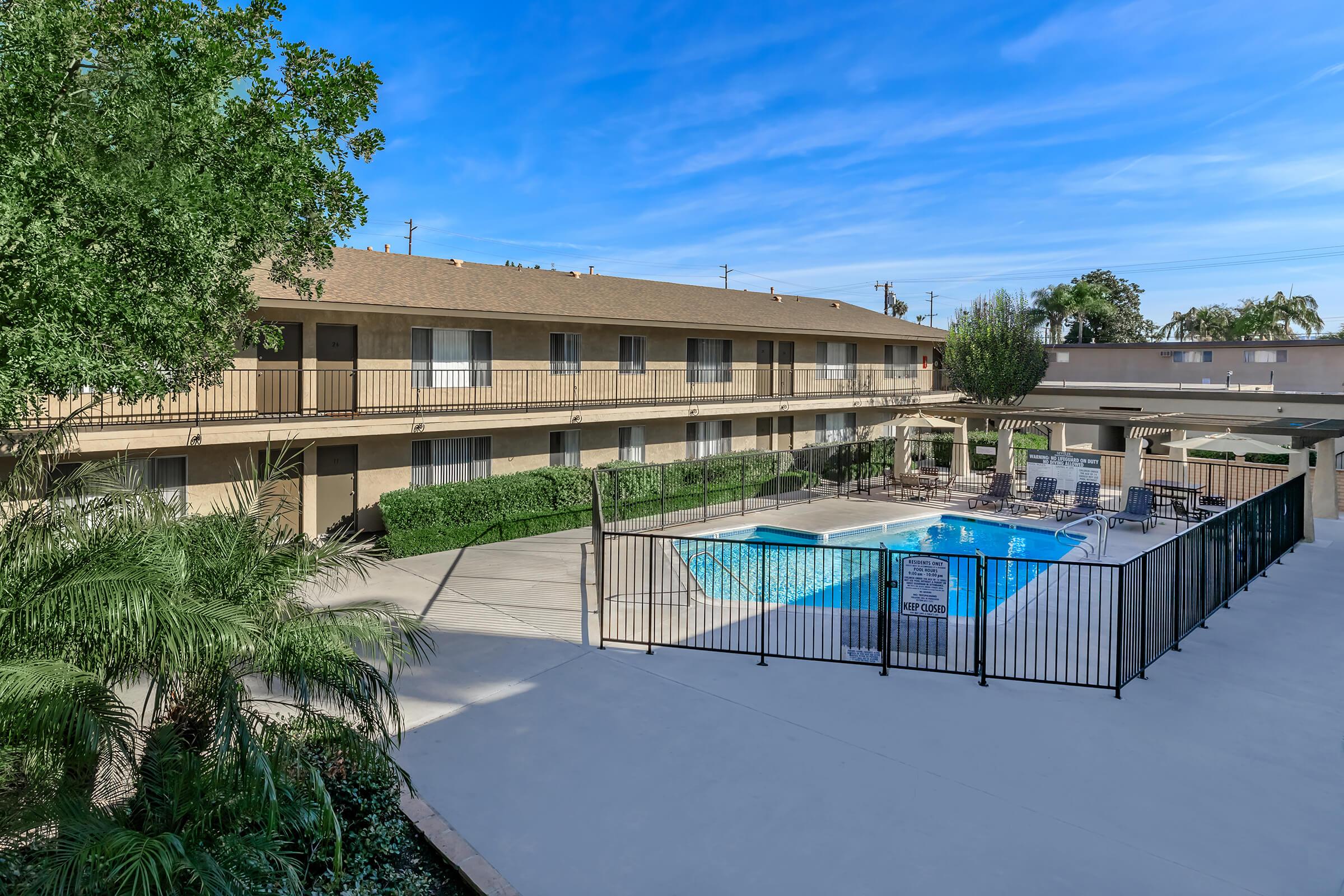 View of a courtyard featuring a swimming pool surrounded by a fence. In the background, a two-story building with multiple rooms and a clear blue sky. Lush greenery and palm trees enhance the outdoor space, creating a welcoming atmosphere for guests. Lounge chairs are set up near the pool.