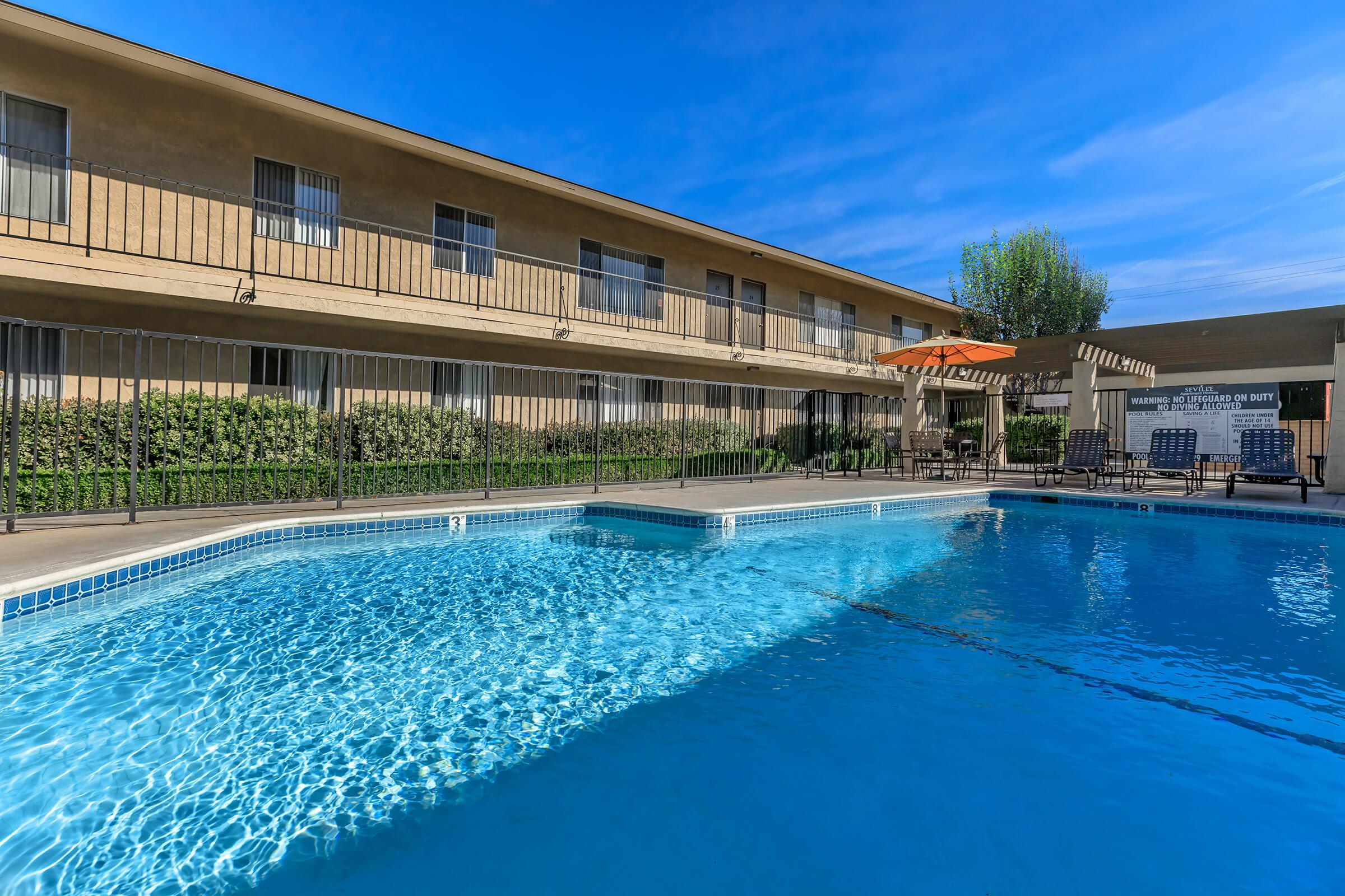 A swimming pool surrounded by a fenced area, with lounge chairs and an umbrella near the water. In the background, a beige building with multiple balconies overlooks the pool area, and green shrubs are visible, creating a serene outdoor setting under a clear blue sky.