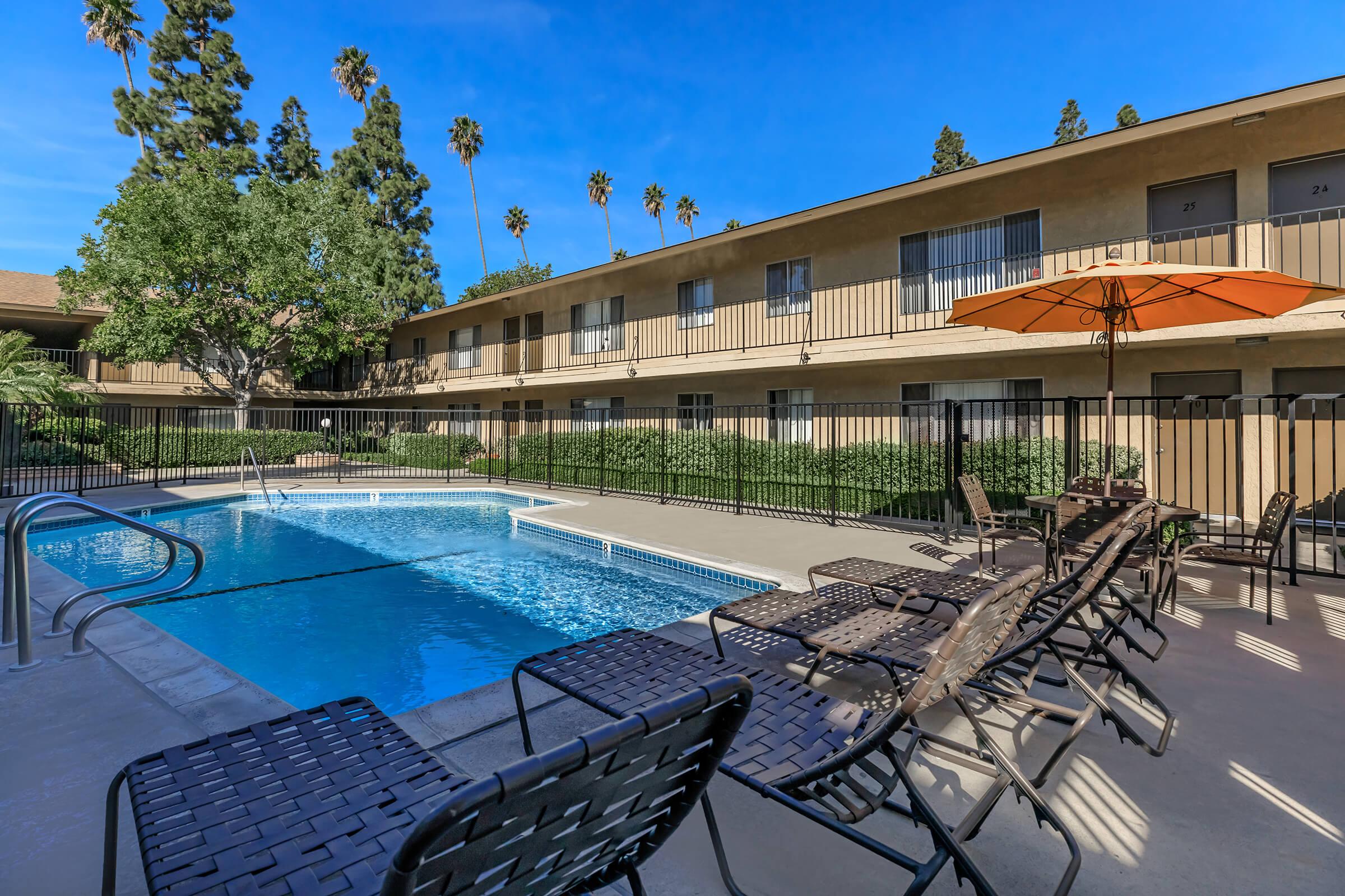 A sunny pool area surrounded by a fence, featuring a swimming pool with clear blue water. Lounge chairs are positioned around the pool, and an orange umbrella provides shade. Tall palm trees and a clear blue sky can be seen in the background, alongside low-rise apartment buildings.