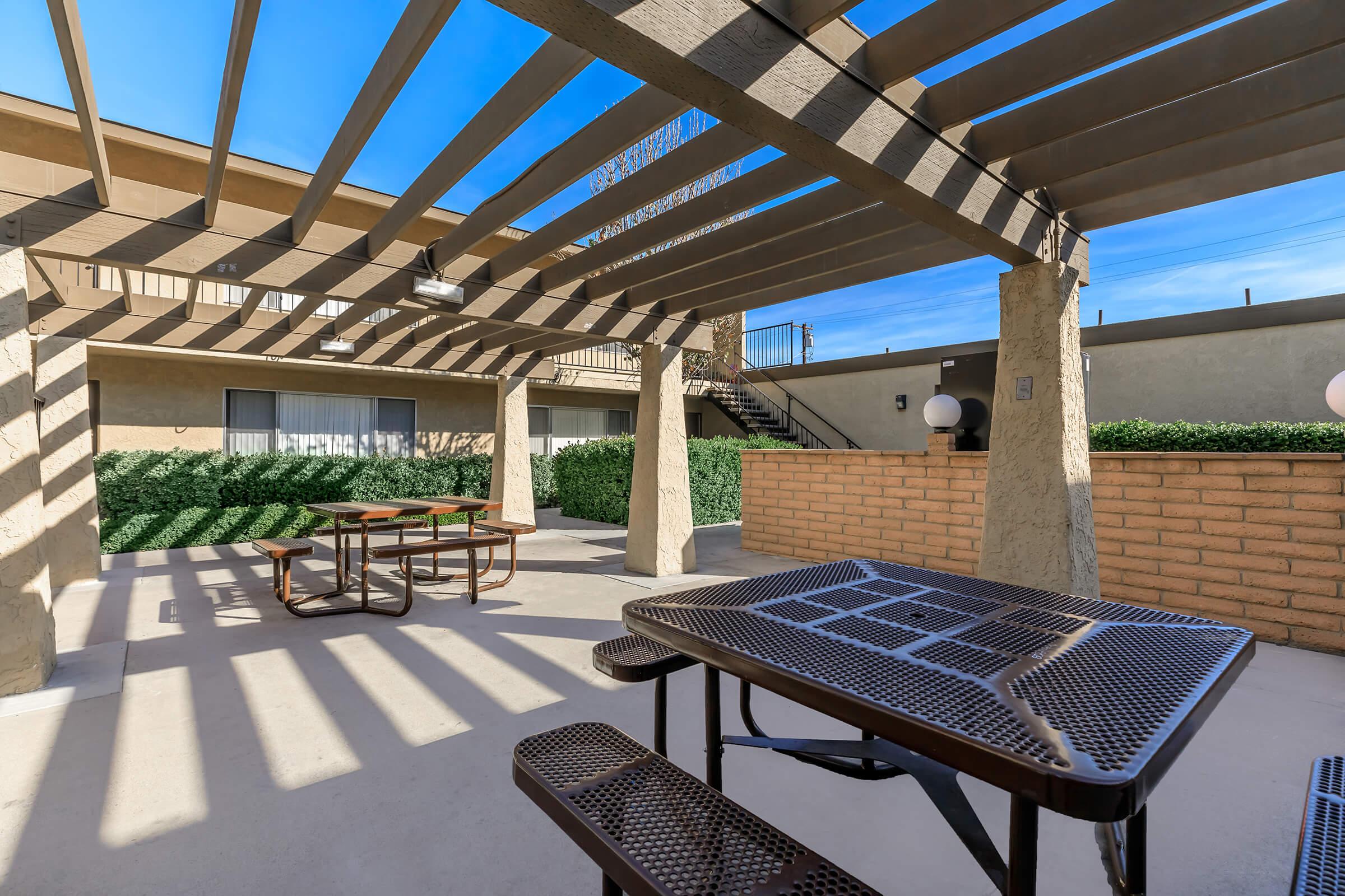 A shaded outdoor patio area featuring metal picnic tables beneath a pergola, with a low brick wall and neatly trimmed hedges in the background. The space is bright and inviting, surrounded by a clear blue sky and well-maintained buildings. Ideal for relaxation or social gatherings.