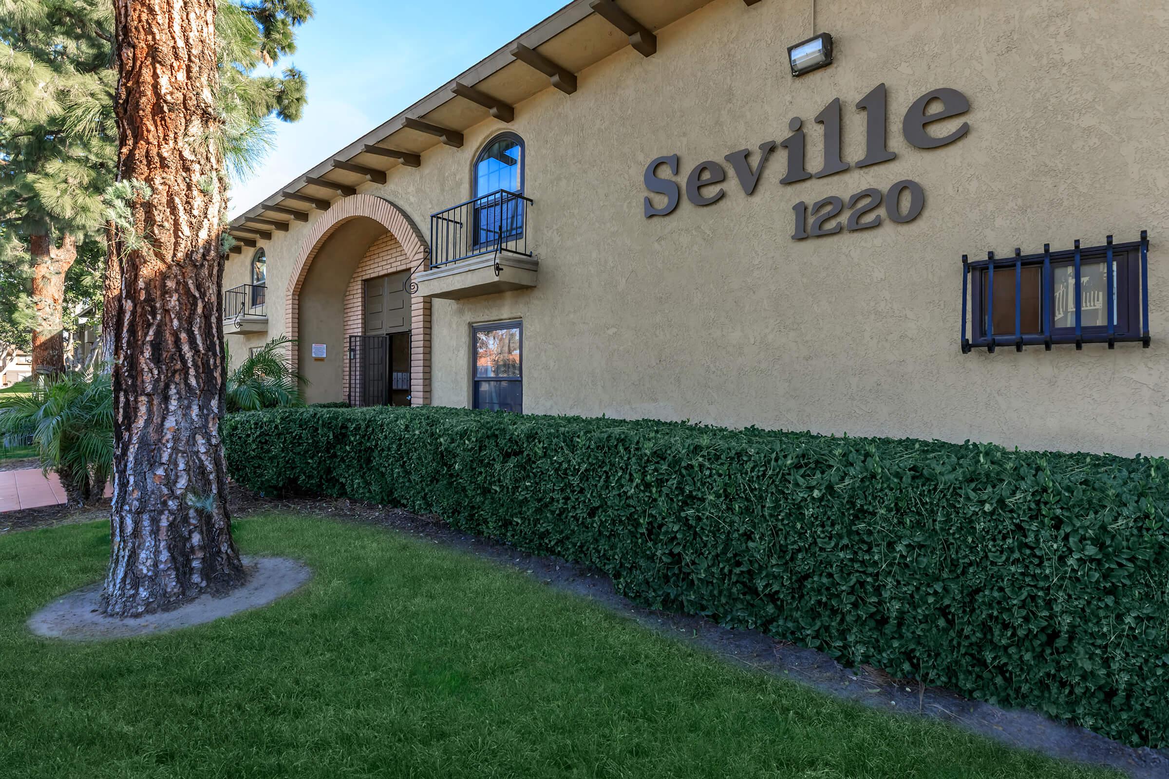 Exterior view of a building labeled "Seville 1220," featuring a textured light-colored wall, large windows, and a landscaped area with manicured hedges and a tall tree. The building design showcases a blend of arches and modern details.