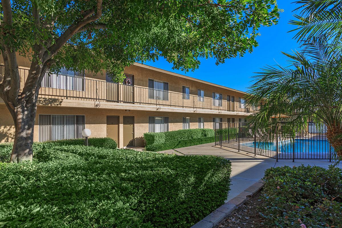 A well-maintained courtyard of a multi-story apartment complex, featuring lush greenery, palm trees, and a swimming pool. The building has balconies and large windows, with a bright blue sky overhead. The area is clean and inviting, ideal for relaxation.