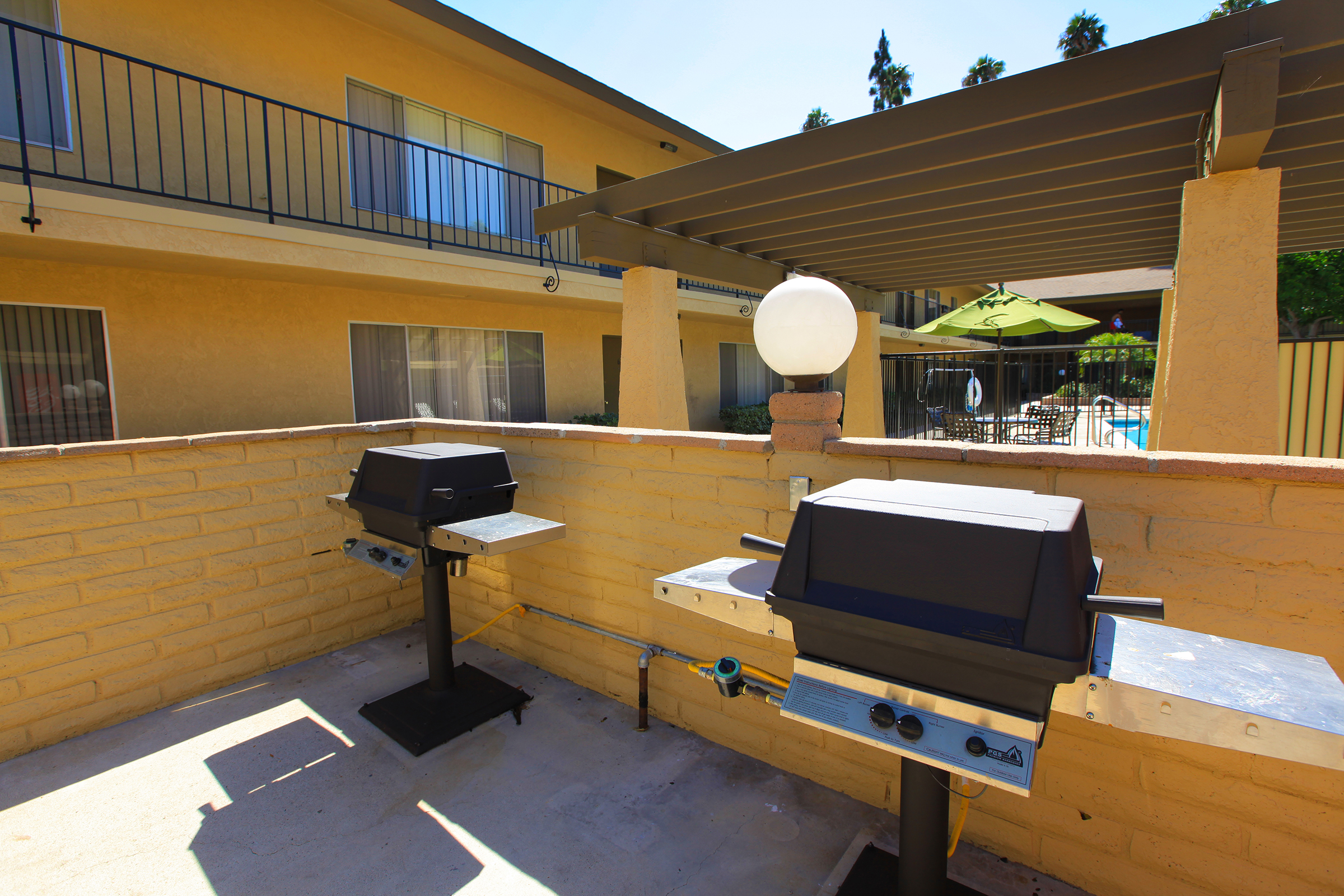 Grill area featuring two outdoor barbecues next to a light post. There is a brick wall surrounding the space, with glimpses of a pool area in the background and some palm trees visible in the sunlight. The setting is bright and inviting for outdoor cooking and gatherings.