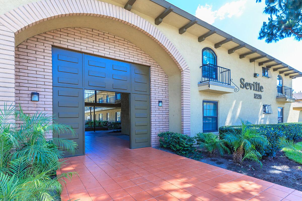 Exterior view of a building entrance featuring large wooden doors with a distinctive arch. The facade is made of beige brick with green accents, surrounded by lush greenery and palm plants. A blue sky is visible above, suggesting a clear day. The building is labeled "Seville" above the entrance.