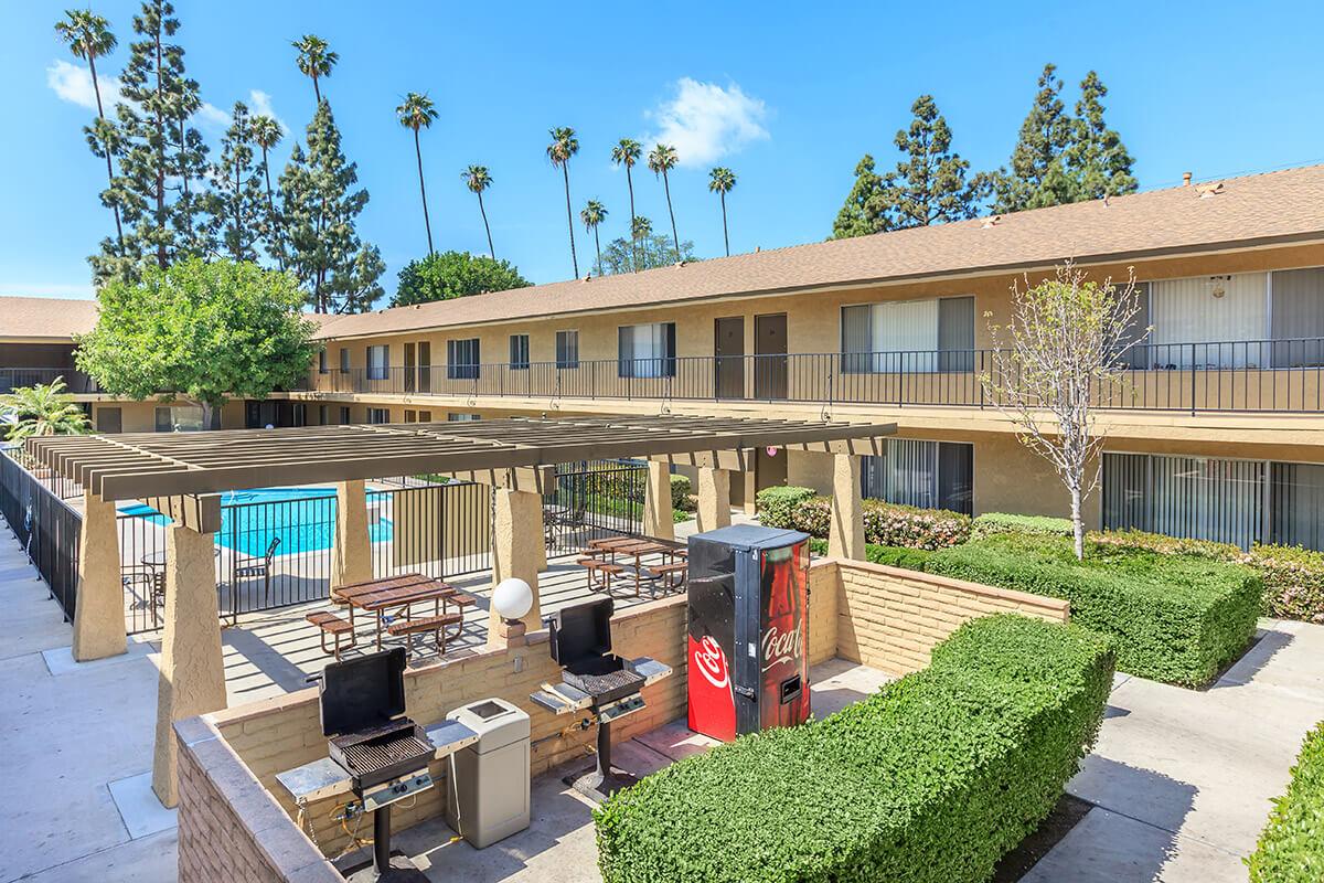 A courtyard view of a two-story motel featuring a swimming pool, green landscaping, and shaded outdoor seating. There are barbecue grills near the seating area, and several palm trees visible against a blue sky. The building has balconies overlooking the courtyard.