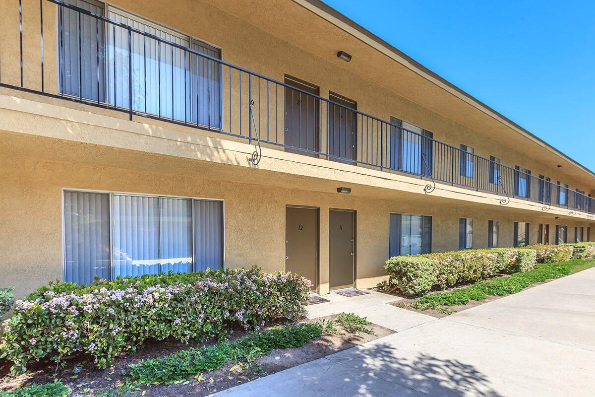 Two-story building with a beige exterior featuring multiple rooms with doors. Each room has a small outdoor balcony and large windows with curtains. Neatly trimmed bushes and plants line the walkway in front of the building. The sky is clear and blue, suggesting a sunny day.