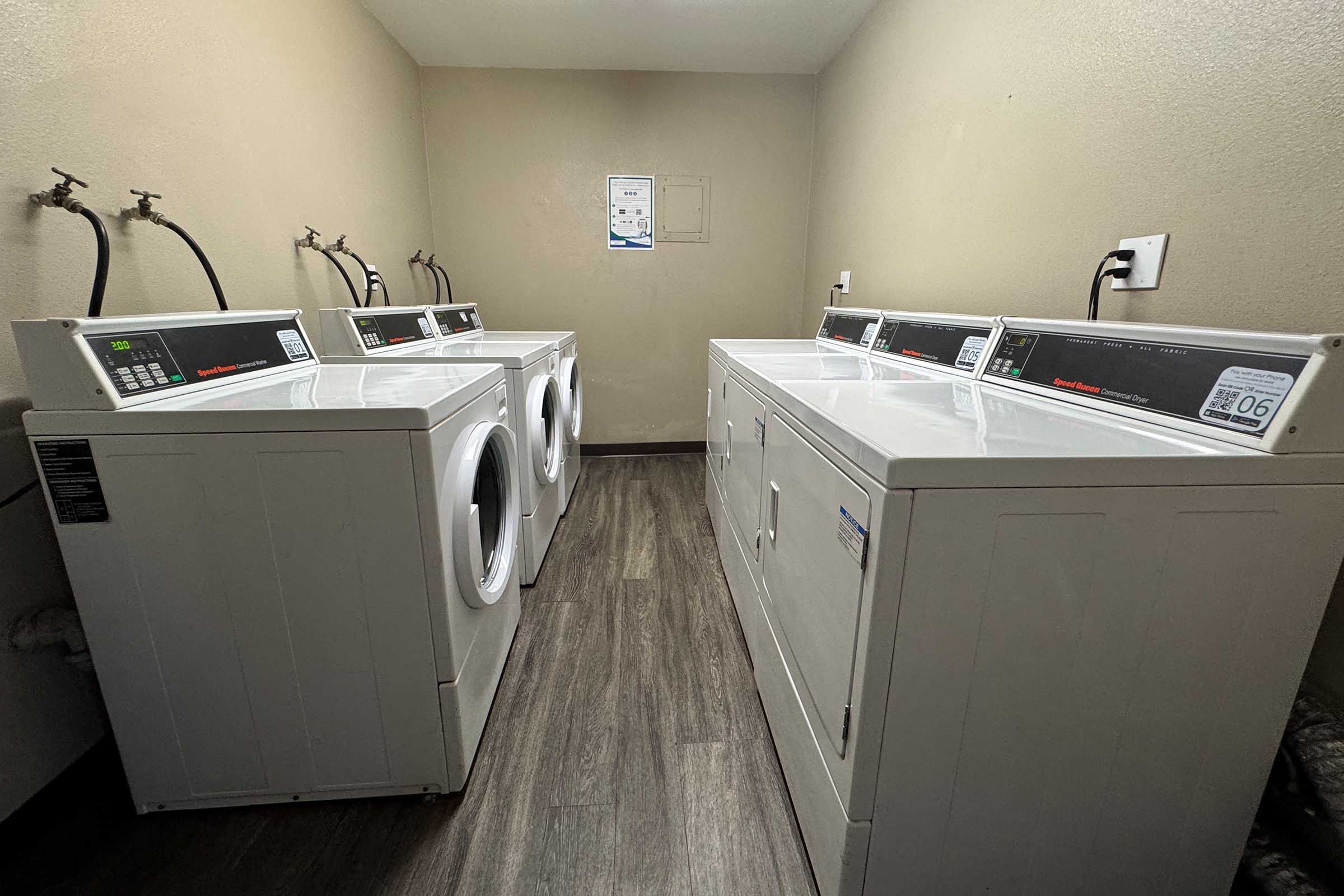 A clean and well-lit laundry room featuring four white washing machines and two white dryers arranged neatly against a light-colored wall. The floor is made of dark wood-like tiles, and there are hooks on the wall for hanging items. A maintenance poster is visible on the wall.