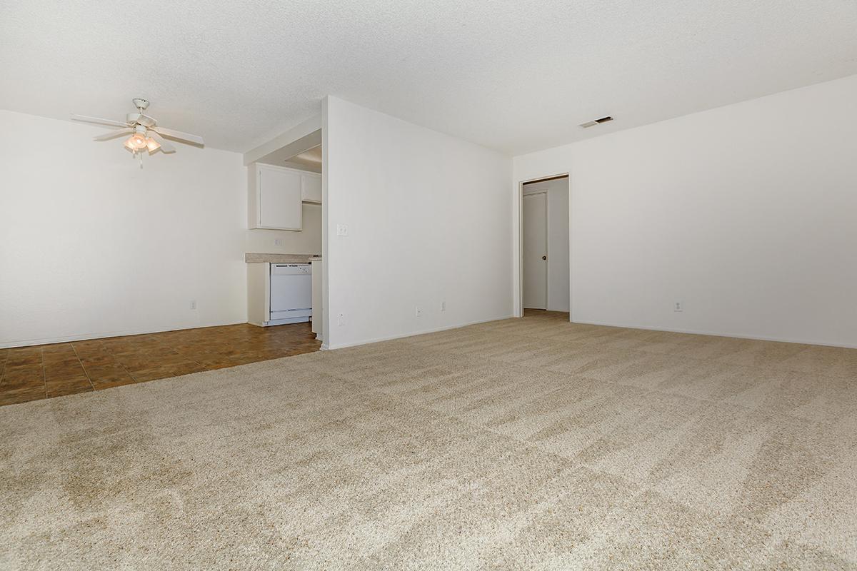 A spacious, empty living room with beige carpet and white walls. A ceiling fan is visible; there's an open doorway leading to a kitchen area in the background equipped with white appliances. The room is well-lit, emphasizing the neutral decor and clean lines, creating a blank canvas for potential furnishings.