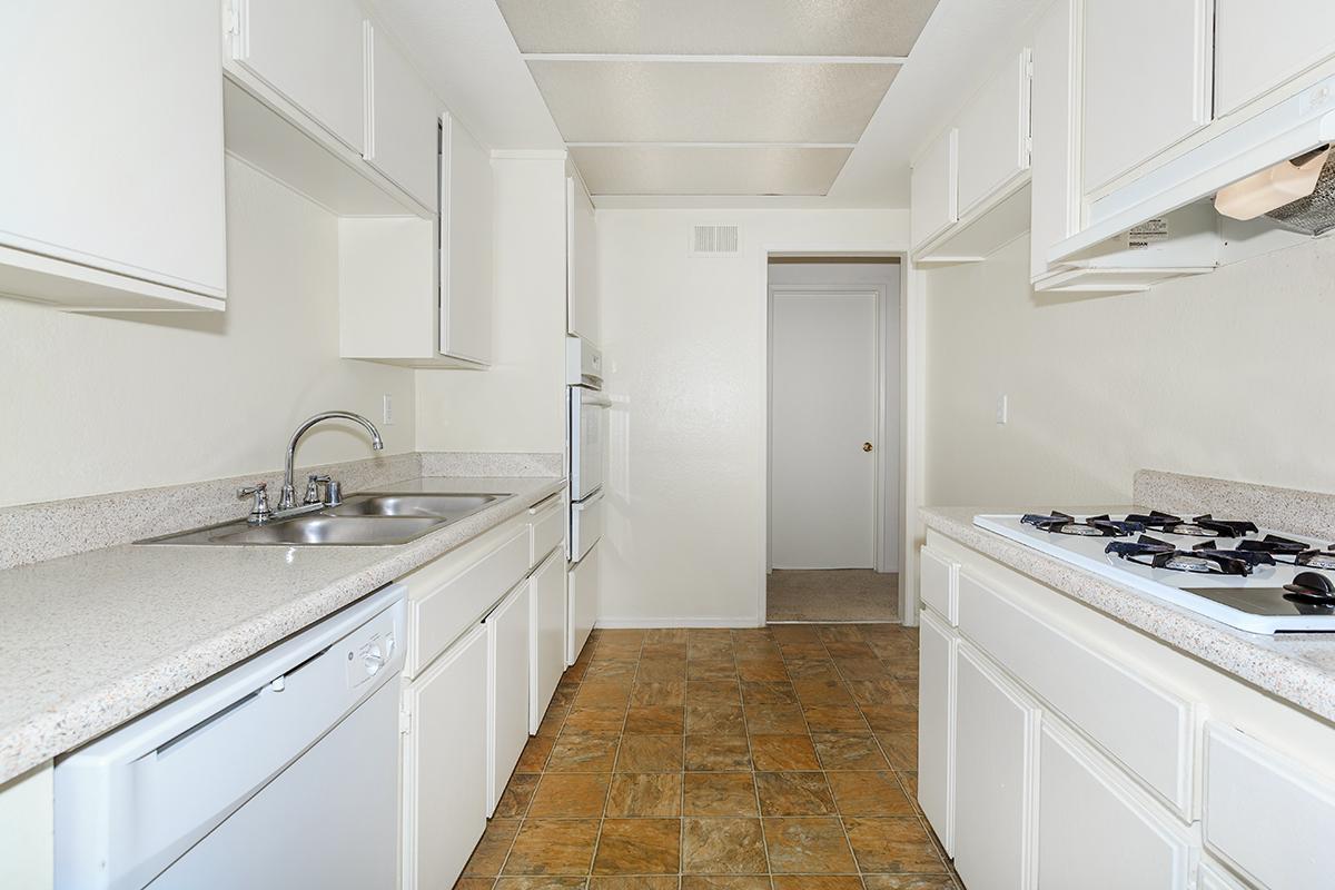 A spacious, modern kitchen featuring white cabinetry, a double sink, and a gas stove. The countertops are light-colored, and the flooring is patterned tiles. The layout includes an adjacent doorway leading to another room, providing a functional and clean design.