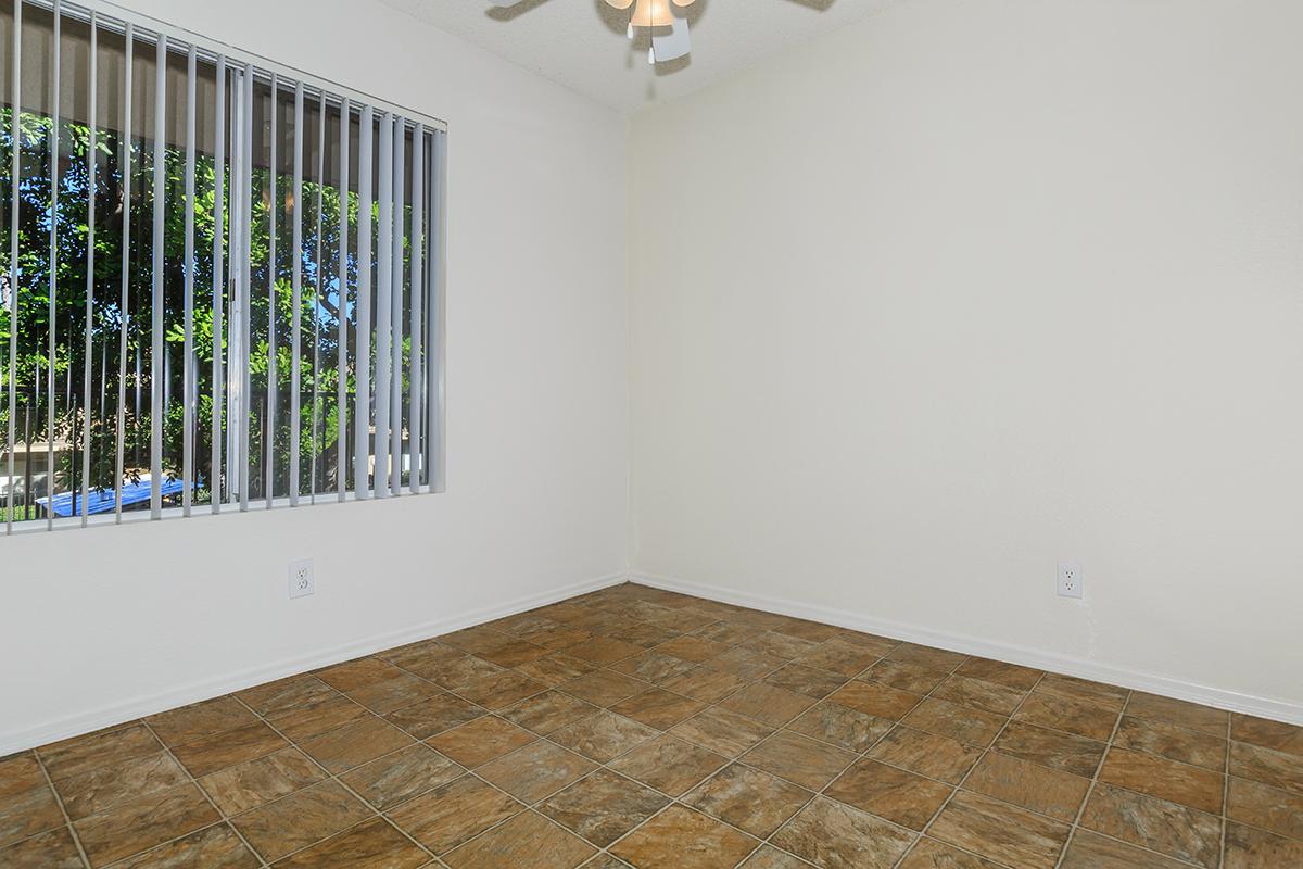 A vacant room with tiled flooring and a white painted wall. There is a window with vertical blinds, allowing natural light to enter, and greenery is visible outside. The ceiling features a light fixture, adding to the brightness of the space.