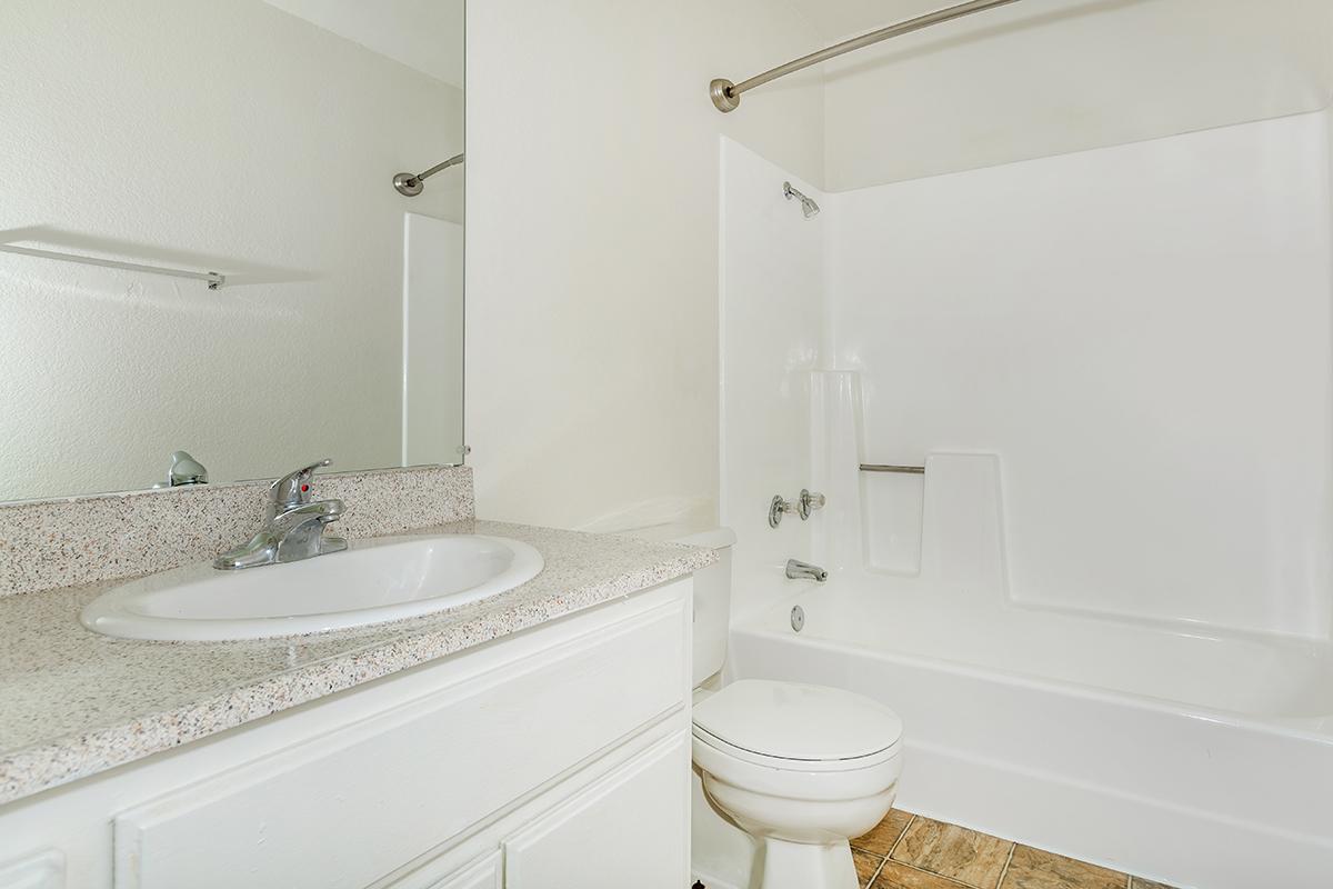 A clean and simple bathroom featuring a white bathtub with a shower, a modern sink with a faucet, a large mirror above the sink, and light-colored walls. The floor is tiled in a neutral tone. The overall aesthetic is bright and minimalist.