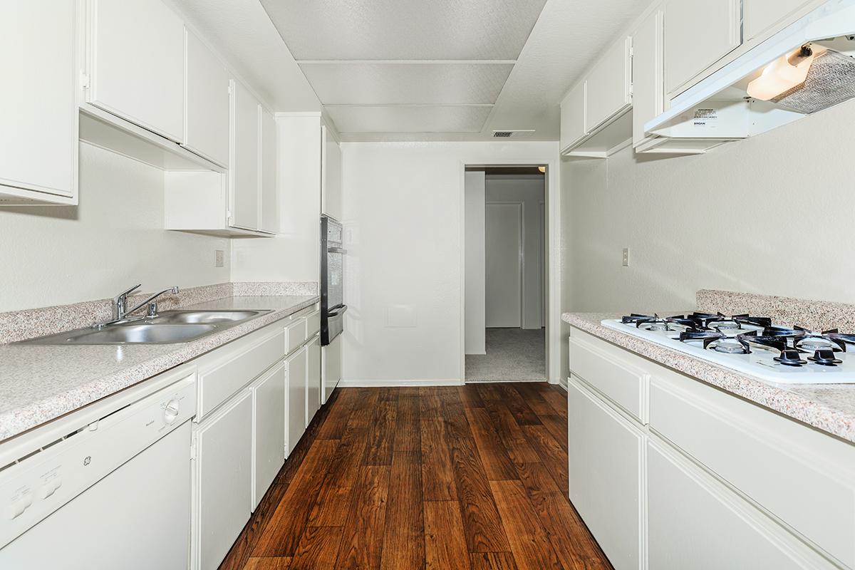 A spacious kitchen featuring light-colored cabinets, a double sink, a gas stove, and a countertop. The floor is covered with dark wooden laminate. The entrance to another room is visible in the background, with neutral-colored walls and carpet.