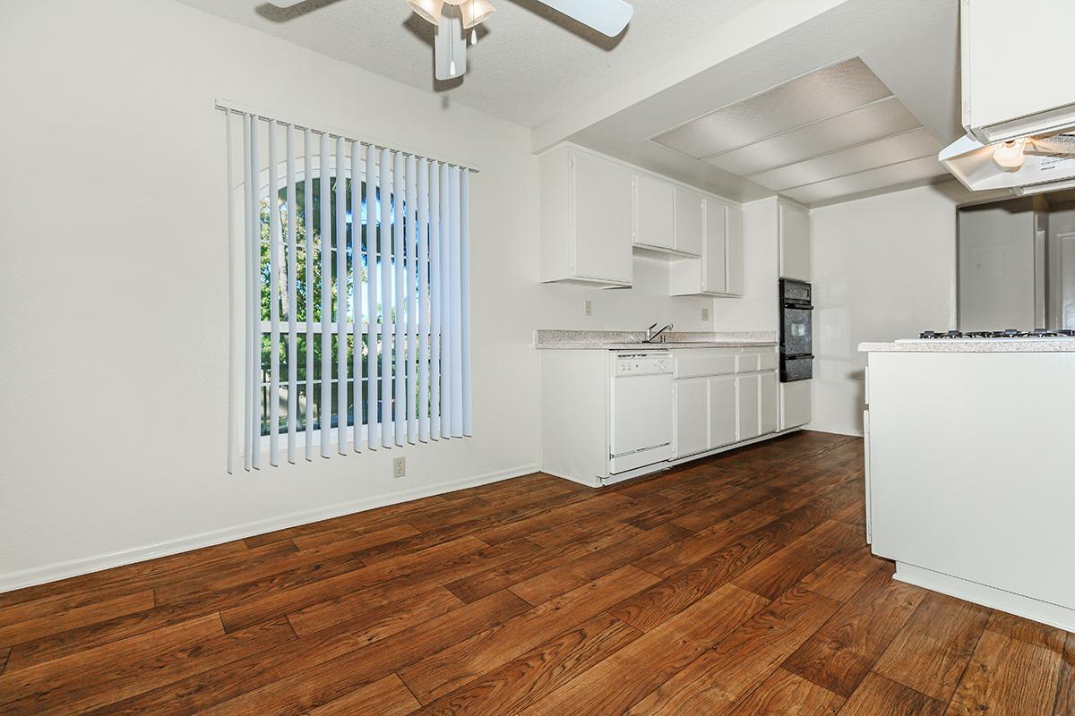 A bright, spacious kitchen featuring white cabinets, a countertop, and a modern black oven. The floor is made of warm-toned wooden planks. A large window with vertical blinds allows natural light to fill the room, and a ceiling fan is visible above. Overall, the space is clean and inviting.