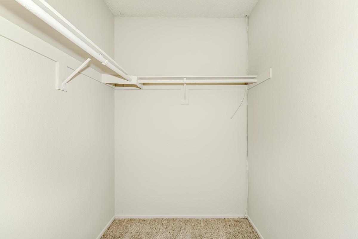 Empty closet with white walls and a beige carpeted floor. A single white rod for hanging clothes is mounted on the wall, with no shelves or additional storage visible. The space is unadorned and minimalist.