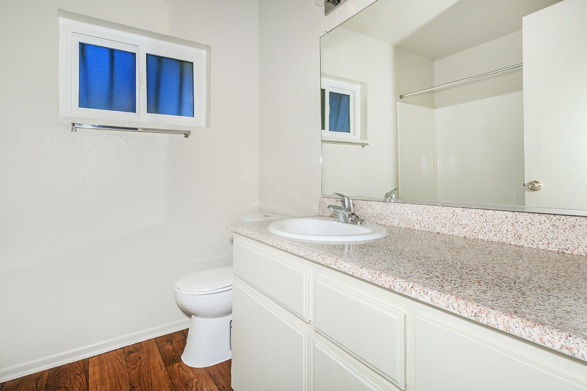A clean, modern bathroom featuring a white countertop with a sink, a large mirror, and a window with blue curtains. The flooring is wooden, and there is a toilet visible in the corner, with light-colored walls adding to the fresh ambiance.