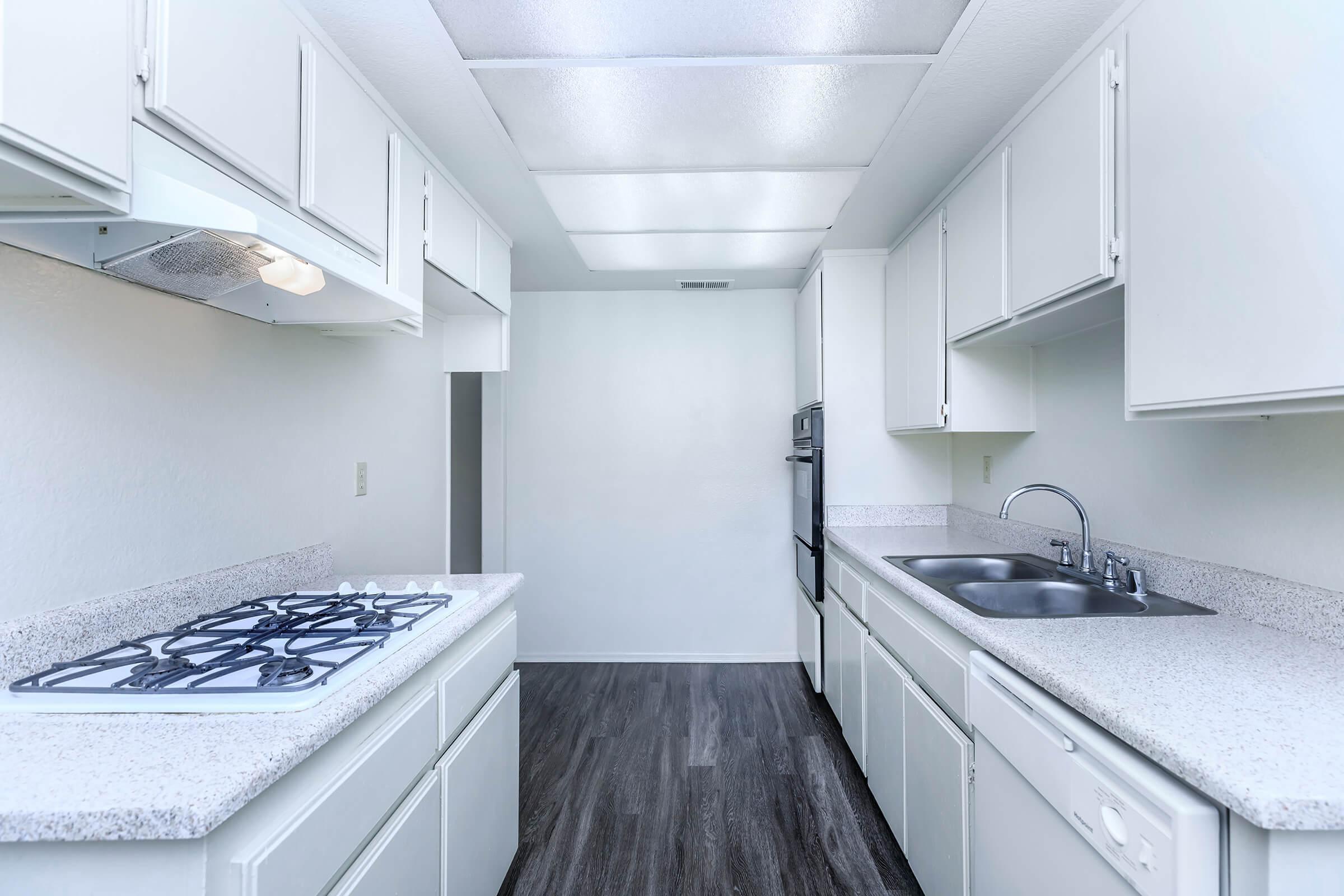 A modern kitchen featuring white cabinetry, a gas stovetop, a double sink, and built-in appliances. The room has a clean, minimalist design with light-colored countertops and a dark wood-like floor, illuminated by overhead fluorescent lighting.