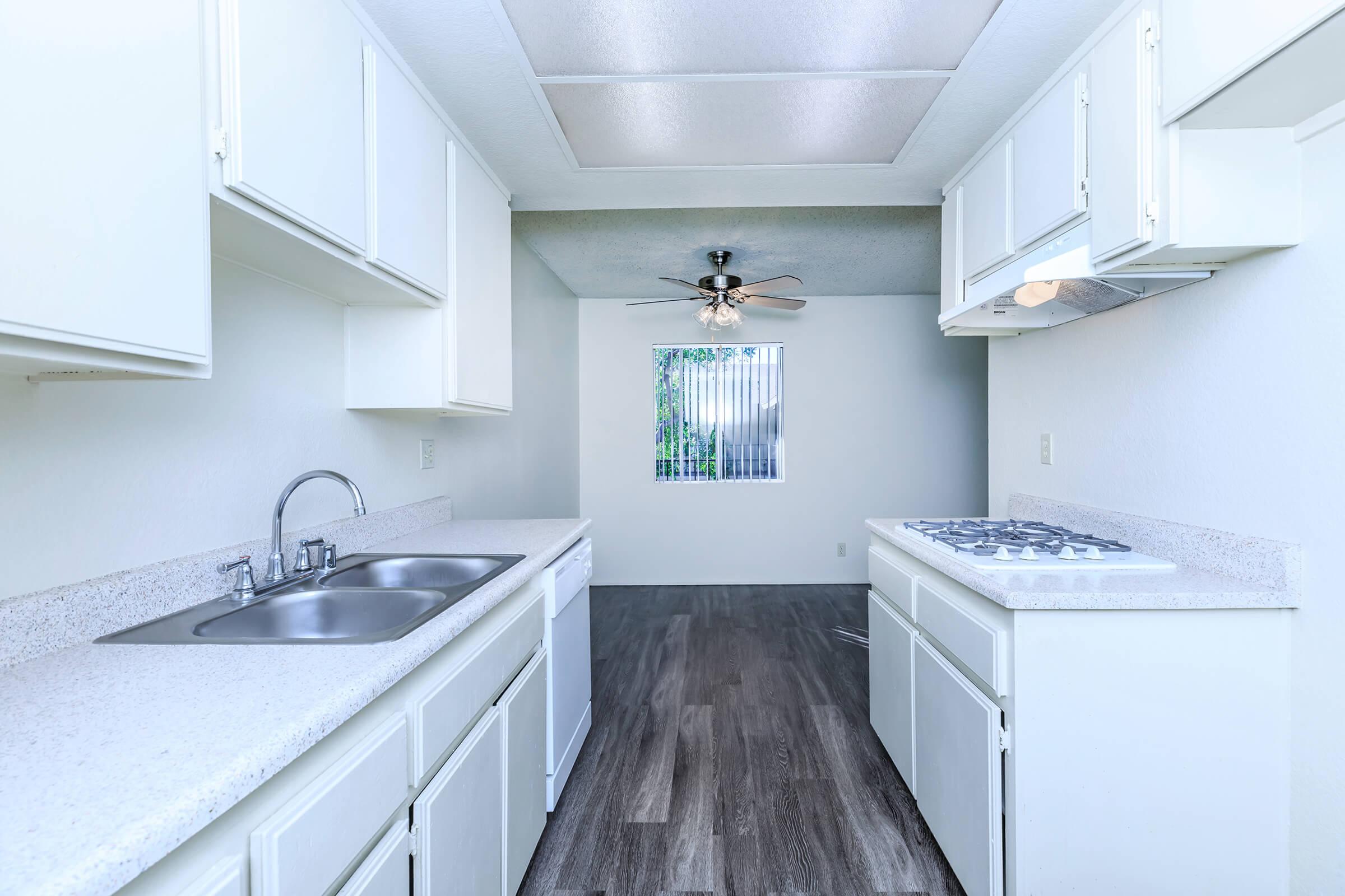 A bright, modern kitchen featuring white cabinetry, a double sink, and a gas stove with a hood. The room has a ceiling fan and a window with vertical blinds, allowing natural light to enter. The flooring is a dark wood-look surface, enhancing the contemporary aesthetic of the space.