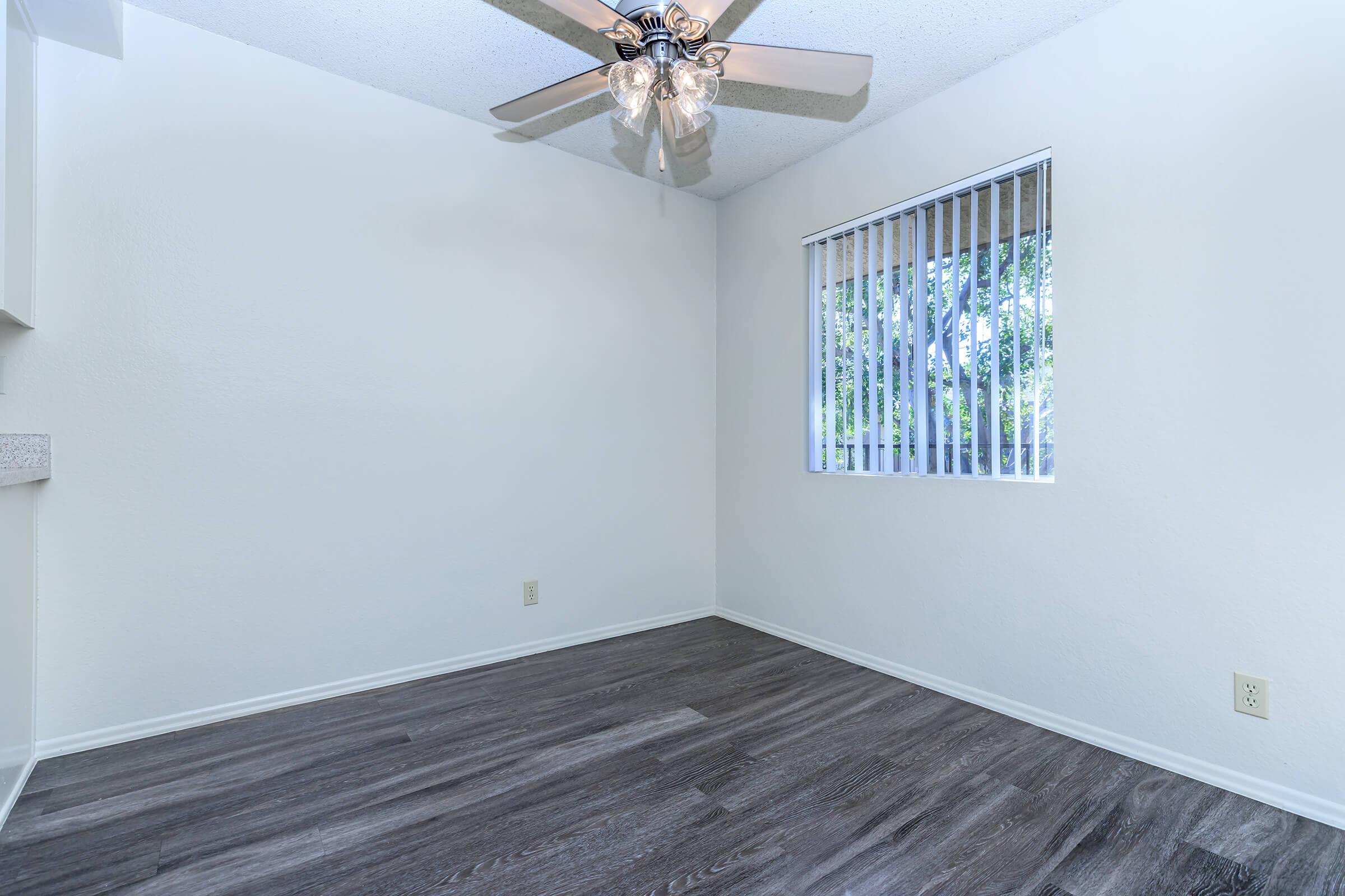 A clean, empty room featuring light hardwood-style flooring, a ceiling fan with five blades, and a window with vertical blinds allowing natural light. The walls are painted white, creating a bright and airy atmosphere.