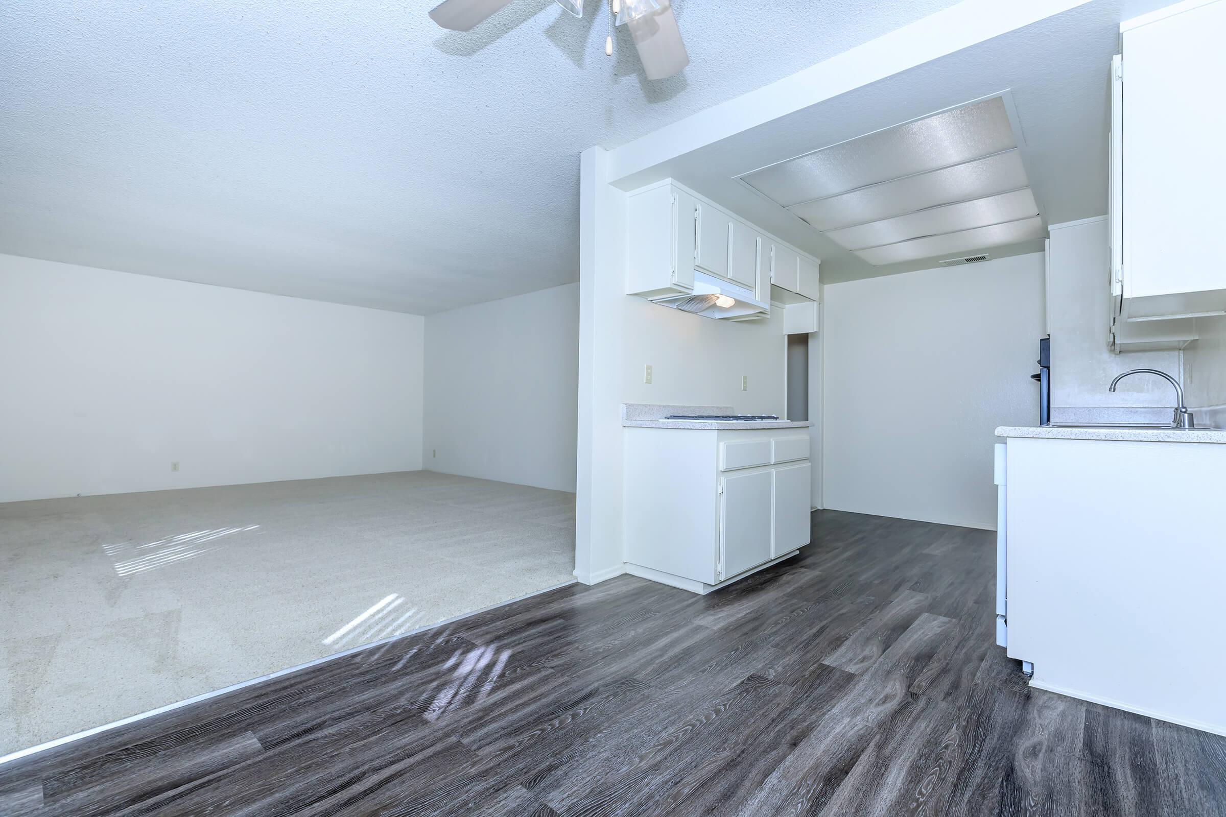 Interior view of an open-concept living space showcasing a kitchen area on the right with white cabinets and a small overhead light. The adjacent living area features light-colored carpeting and white walls, creating a bright and airy atmosphere. There's a ceiling fan visible above.