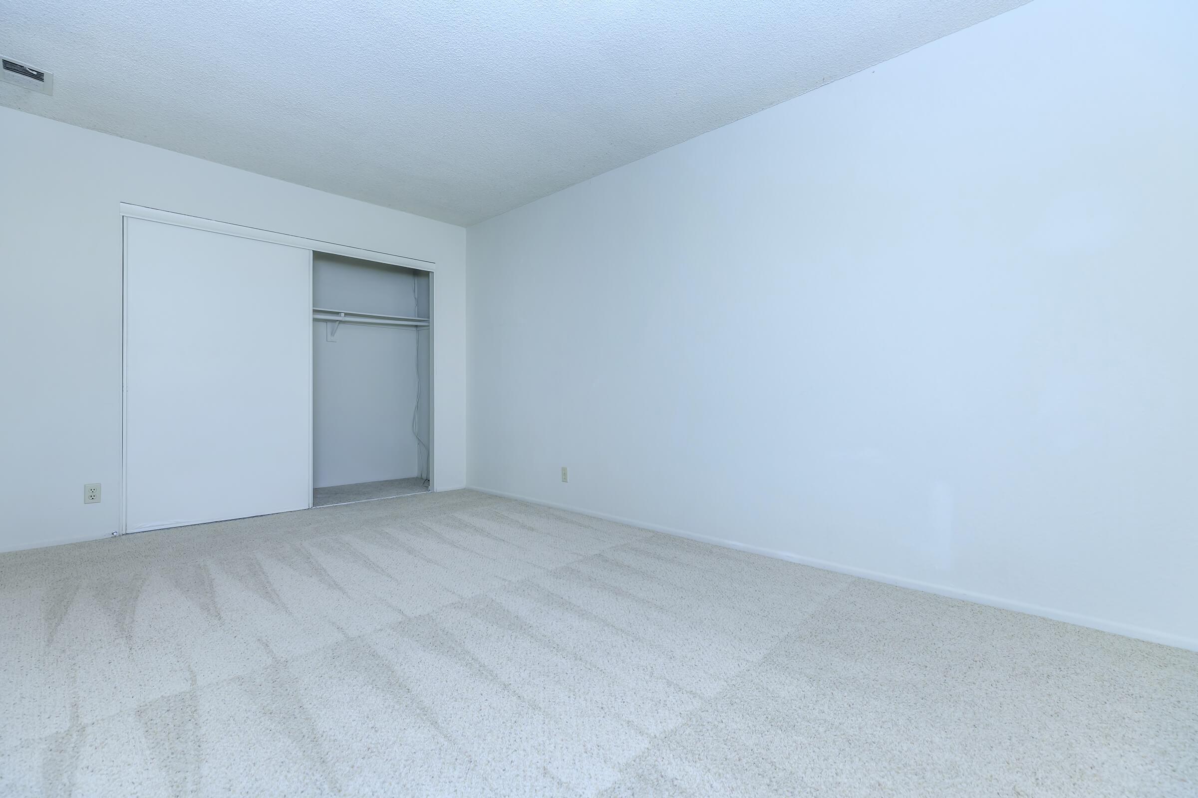 Empty room with light-colored carpet and white walls. A closet with sliding doors is visible on the left side. The space is well-lit, creating a bright and neutral atmosphere, ideal for various uses or decorations.