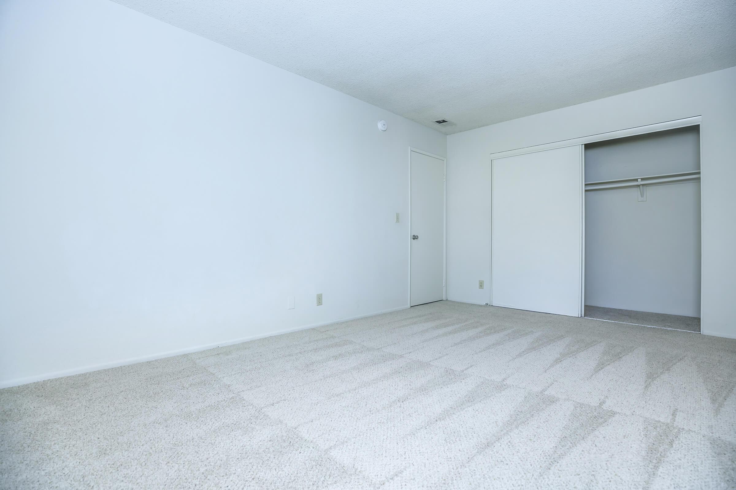 Empty room with light-colored walls and a carpeted floor. There is a white door on one side and a closet with sliding doors. The room has no furniture or decorations, creating a spacious and clean appearance.