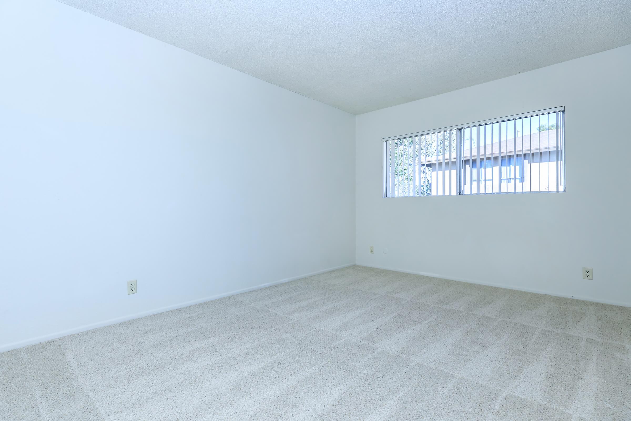 A spacious, empty room with light-colored walls and a soft, beige carpet. There is a window with metal bars on one side, allowing natural light to enter the space. The room appears clean and ready for furnishings, offering a blank canvas for decoration or use.