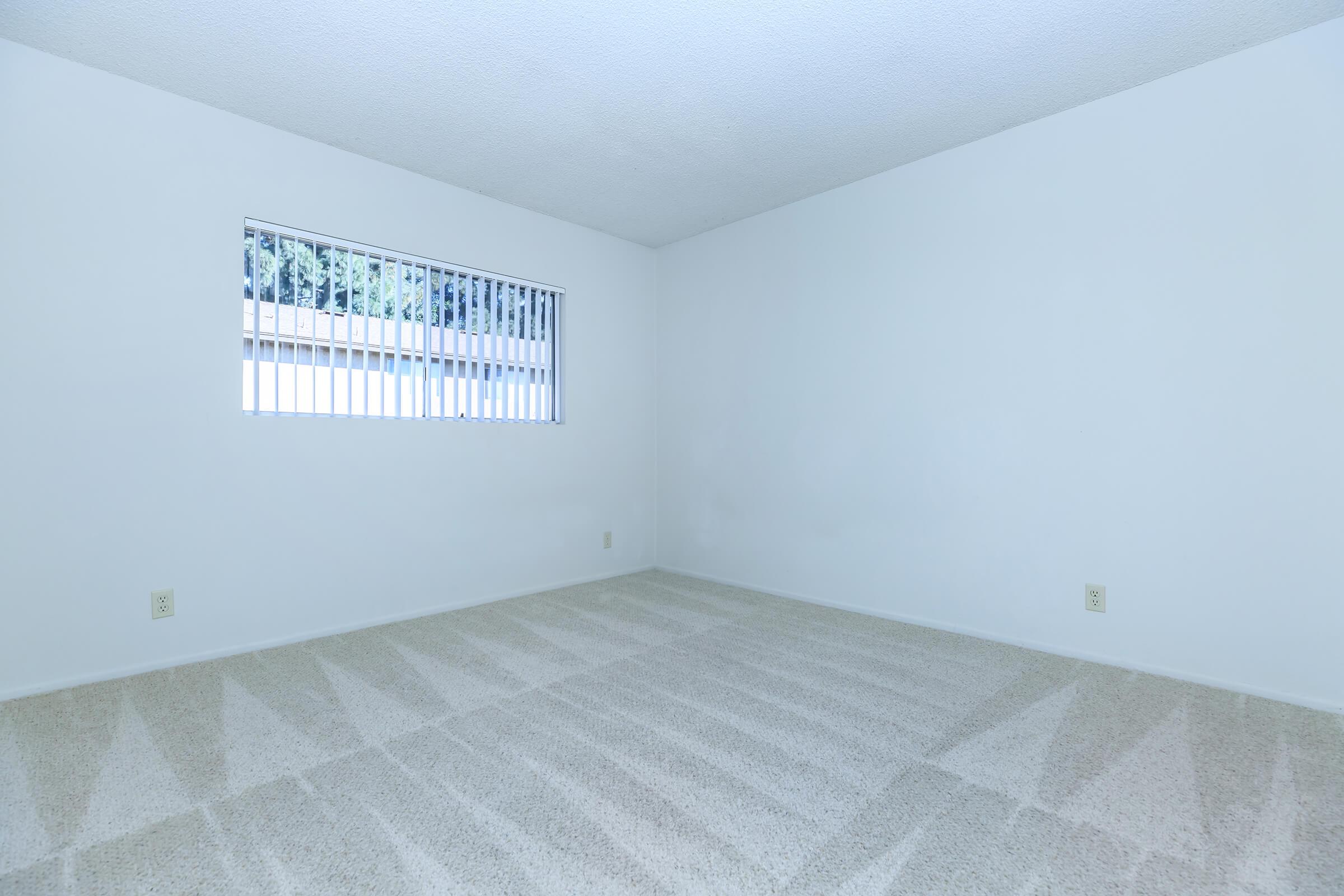 A sparsely furnished room with light-colored walls and beige carpet. The only window features horizontal blinds, allowing soft natural light to enter. The space is empty, providing a blank canvas for decoration and furniture arrangement.