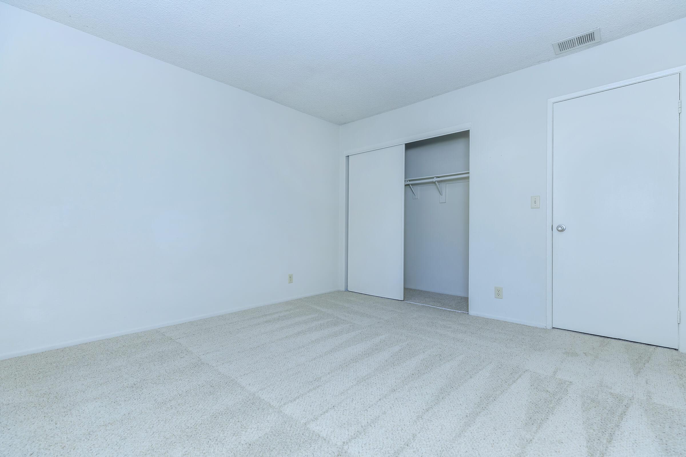 A spacious, empty room with white walls and light-colored carpet. There is a closet with a sliding door on the left side, and a closed door on the right. The room has a bright and clean appearance, ideal for furnishing or decorating. Natural light brightens the space.