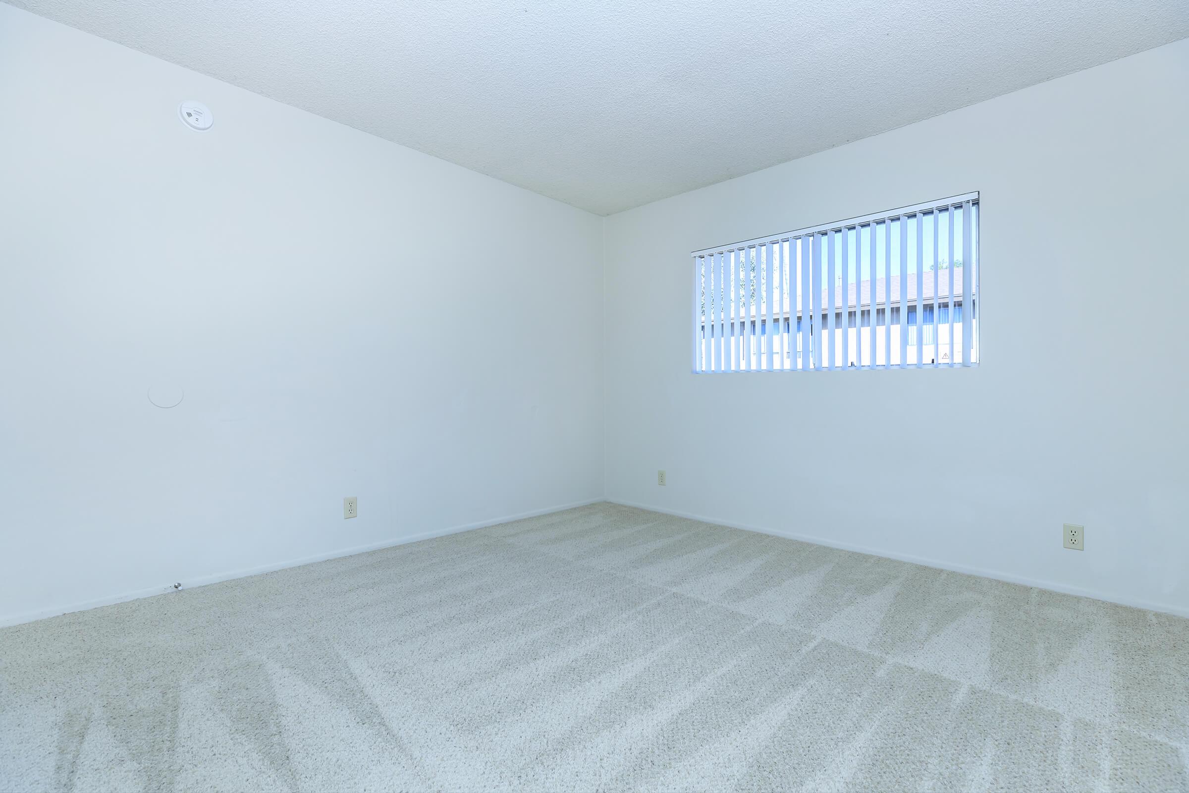 Empty room with light-colored walls and carpeted flooring. A single window with vertical blinds allows natural light to enter. The space appears clean and minimalist, with no furniture or decorations. There are electrical outlets on the walls.
