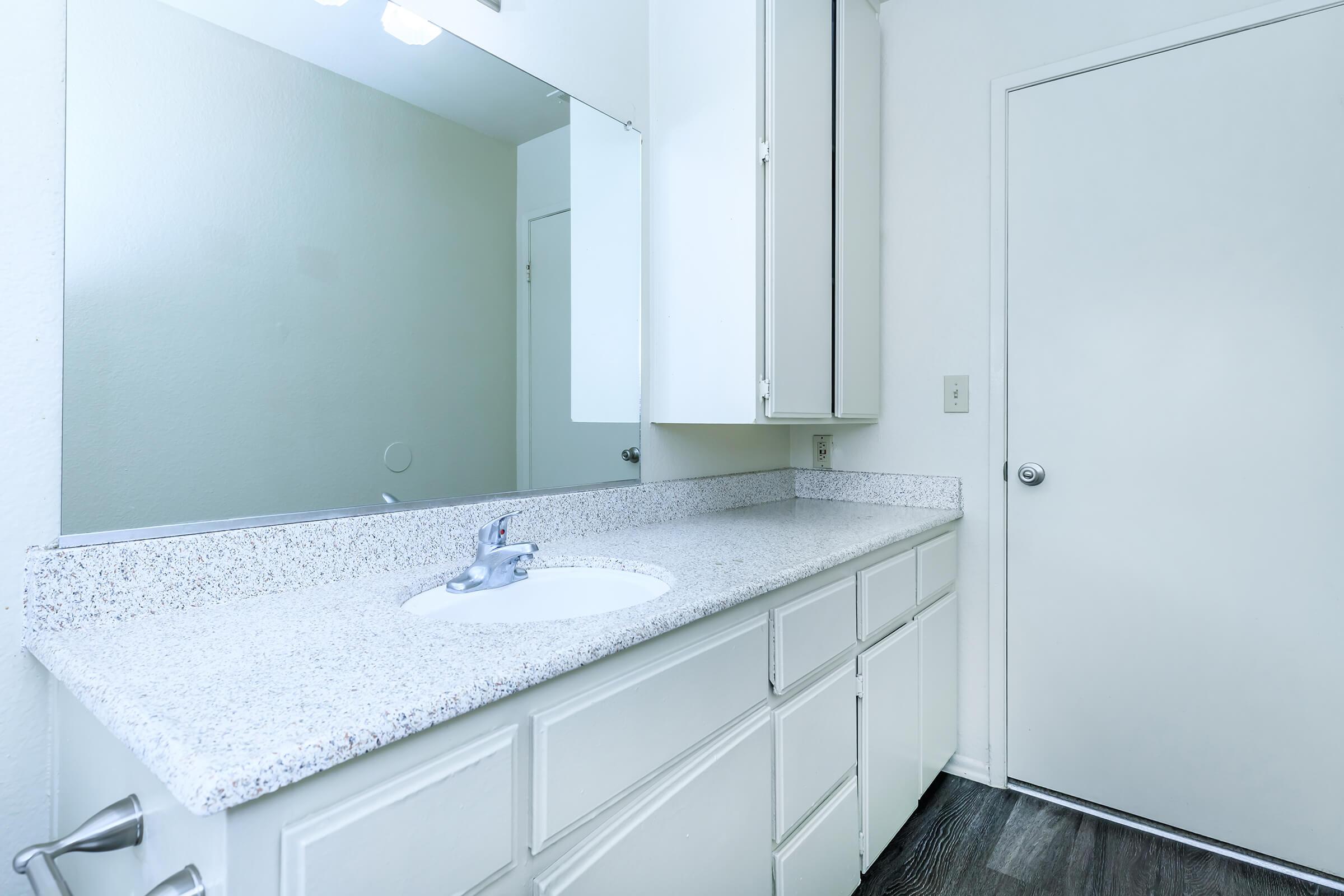 A clean bathroom featuring a light-colored granite countertop with a sink, large mirror above, white cabinetry, and a door. The walls are painted a neutral color, and the flooring appears dark wood. Natural light is coming from an unseen source, enhancing the bright atmosphere.