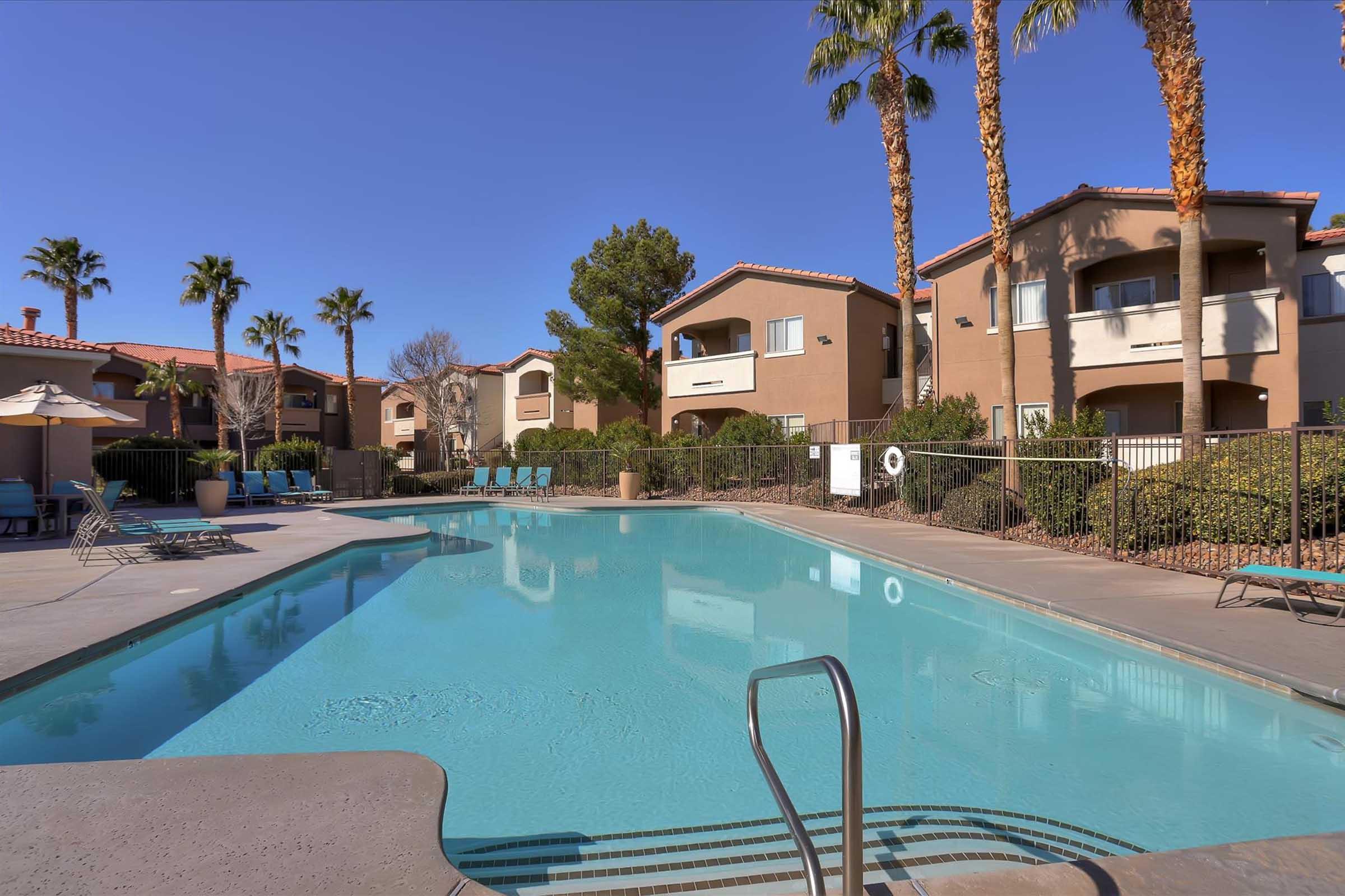 Swimming pool surrounded by palm trees and apartment buildings. The water is clear and inviting with a pool ladder visible. Lounge chairs are arranged nearby, and a few umbrellas provide shade. The sky is clear and blue, creating a relaxing outdoor atmosphere.