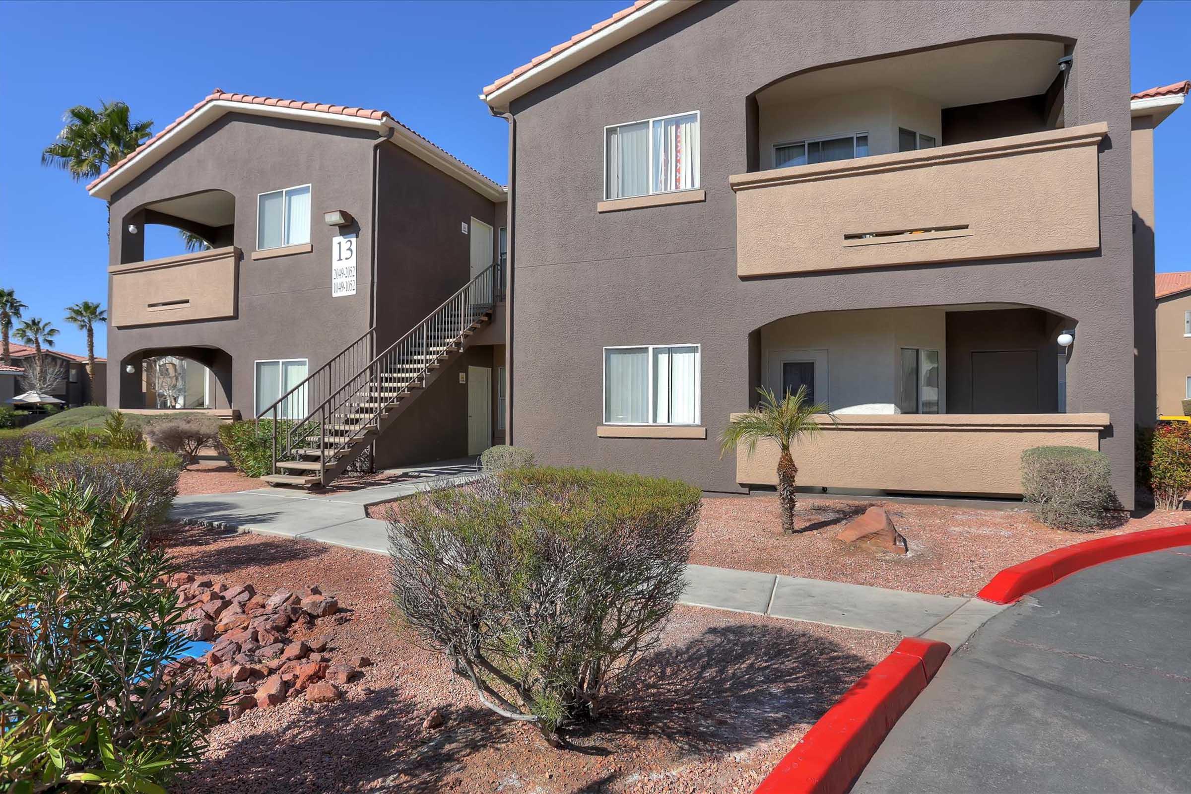 Two-story apartment building with a brown exterior and red accents. The entrance features a set of stairs leading to the second floor. The landscape includes small shrubs and palm trees, with a clear pathway and a portion of a red-painted curb visible. Bright sunlight casts soft shadows around the scene.