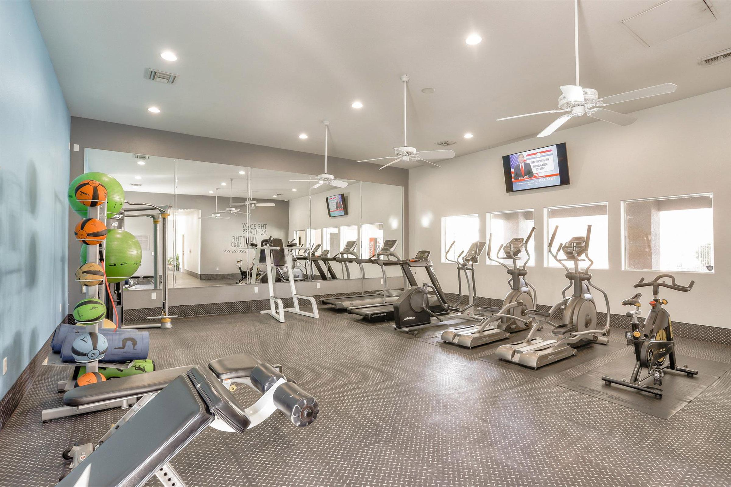 A well-lit gym area featuring various exercise equipment, including treadmills, ellipticals, and stationary bikes. The space has large mirrors, a blue accent wall, ceiling fans, and a wall-mounted TV, creating a modern and inviting workout environment.