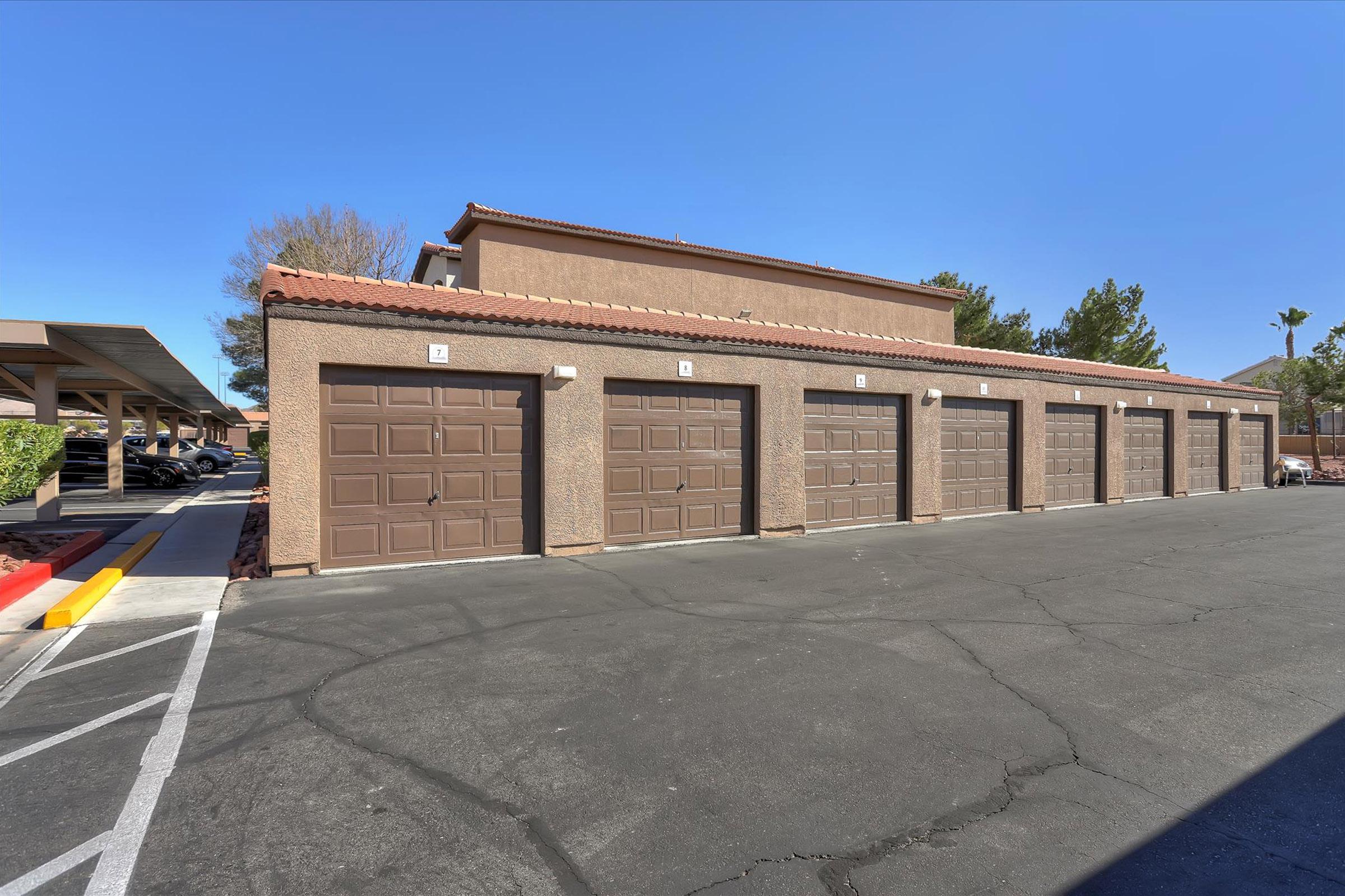A row of closed garage doors in a well-maintained parking area, with a clear blue sky above. The garages are part of a larger building in the background, surrounded by trees and parked cars. The asphalt surface of the parking lot is visible in the foreground.