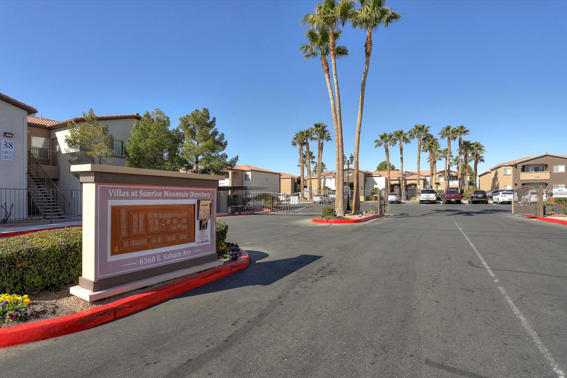 View of a residential community entrance featuring a sign for "Villas at Sunrise Mountain." The scene includes palm trees lining the street, a clear sky in the background, and well-maintained buildings on both sides of the road. The area is landscaped with flowers and greenery, creating an inviting atmosphere.