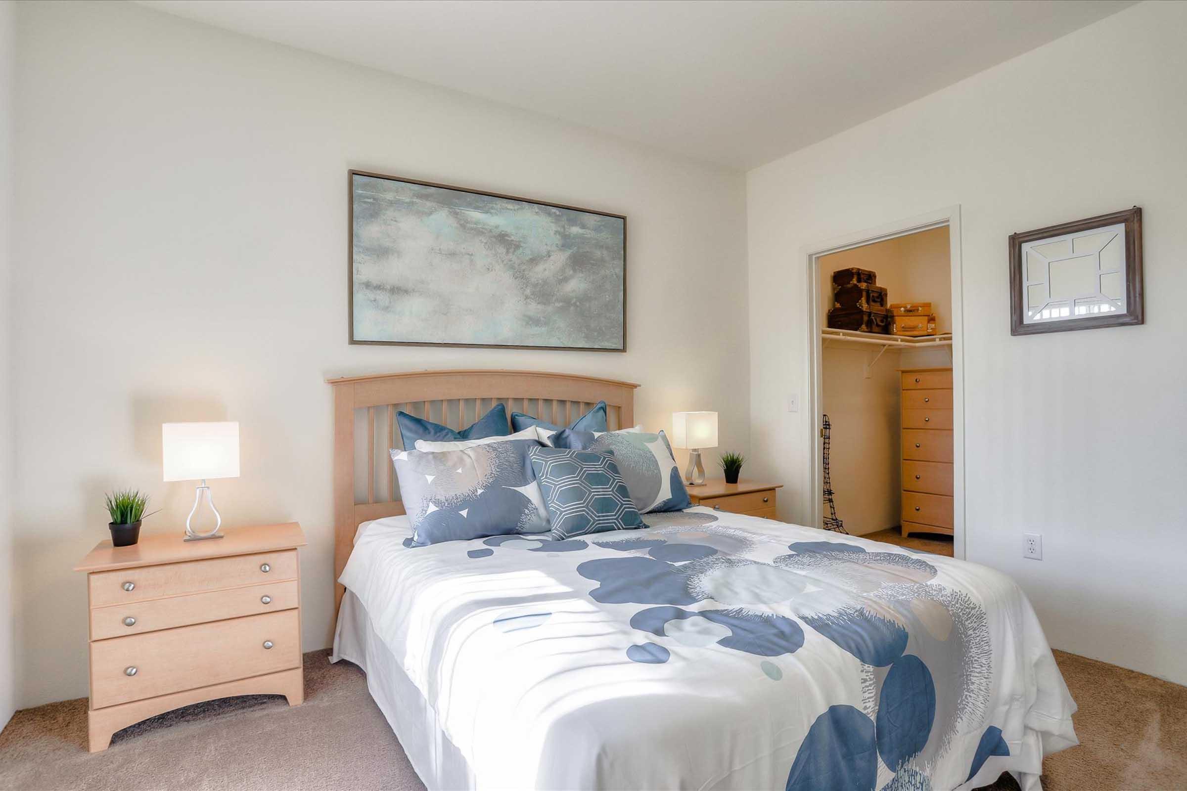 A well-decorated bedroom featuring a queen-sized bed with a white and blue floral comforter, matching pillows, and bedside tables with lamps. The walls are painted white, and a large abstract painting hangs above the bed. An open closet is visible, showing a dresser and storage space.